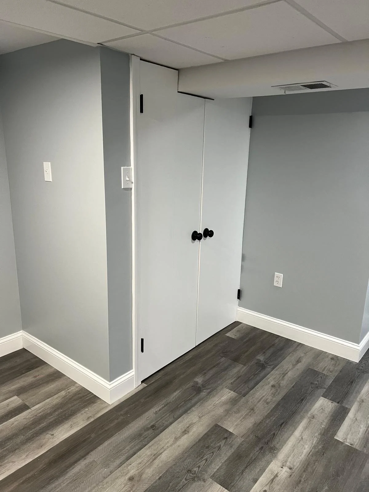 Corner of a room with light gray walls, a ceiling with white tiles, a white closet with black knobs, and wood-look vinyl flooring in shades of gray and brown.
