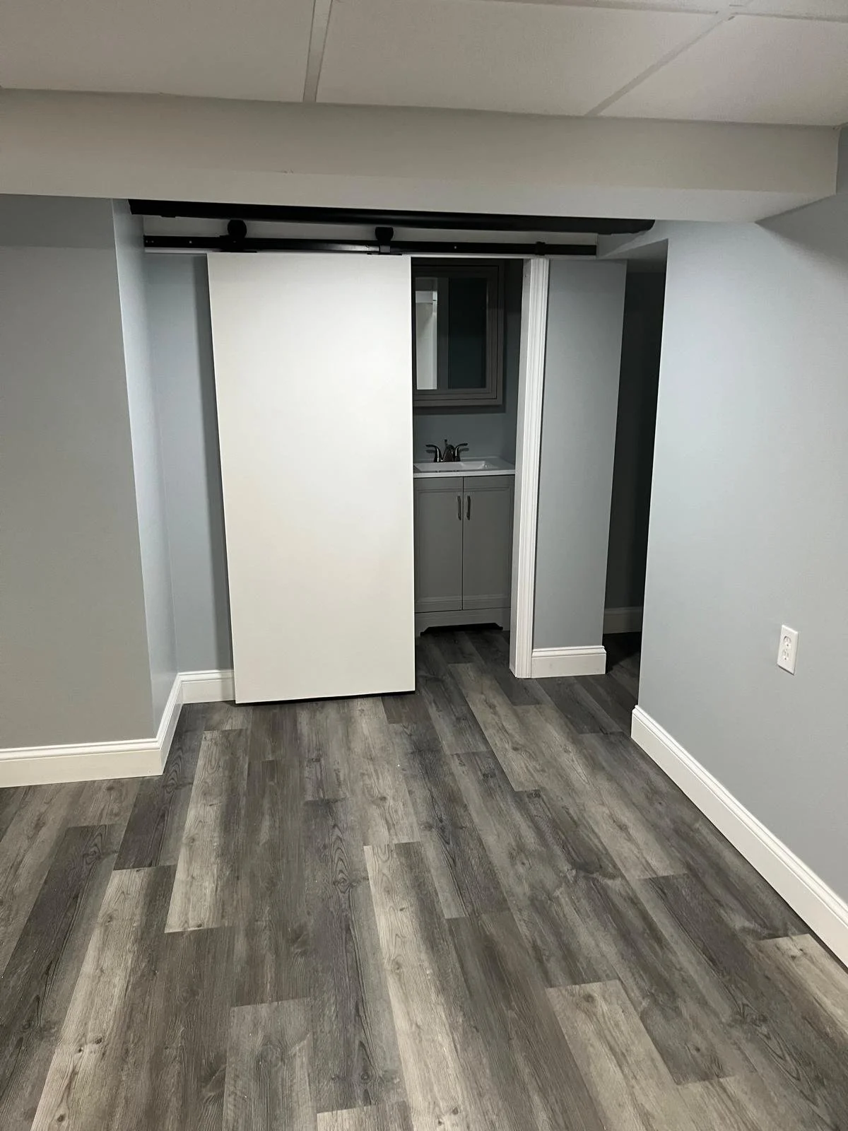 Empty room with gray walls and wood-look vinyl flooring, featuring a small bathroom with a vanity and mirror behind a partially open white sliding door.