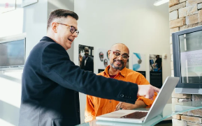 Two men collaborate at a desk with a computer, smiling and shaking hands in a modern office.
