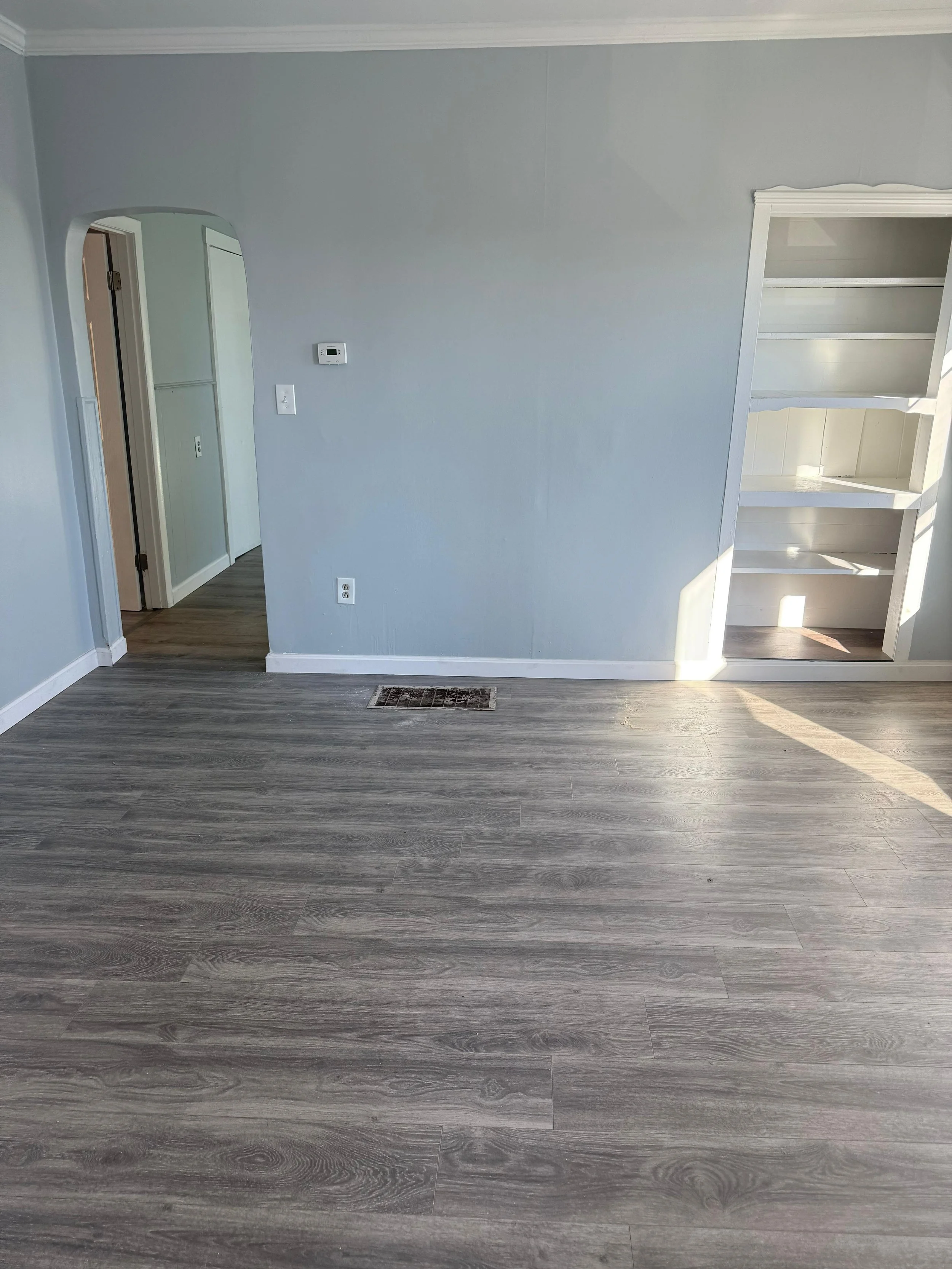 Empty room with grey wood flooring, pale blue walls, built-in white shelves, and a small wall vent near the floor. There is an arched doorway leading to another room and a thermostat on the wall.