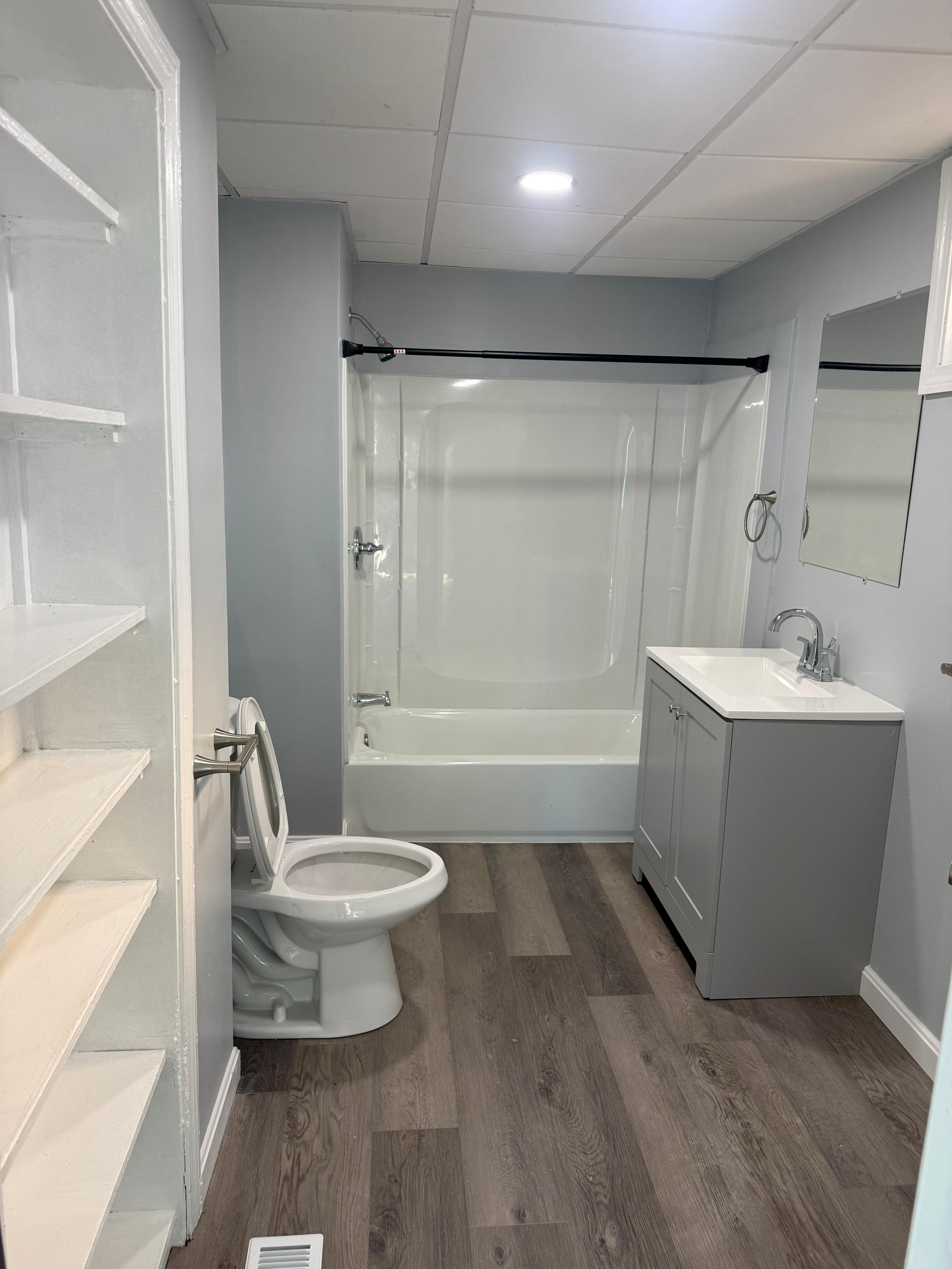 A small bathroom with a shower-tub combination, a toilet to the left, and a small vanity with a sink and mirror to the right. The walls are painted light gray, and the floor has wood-like planks.