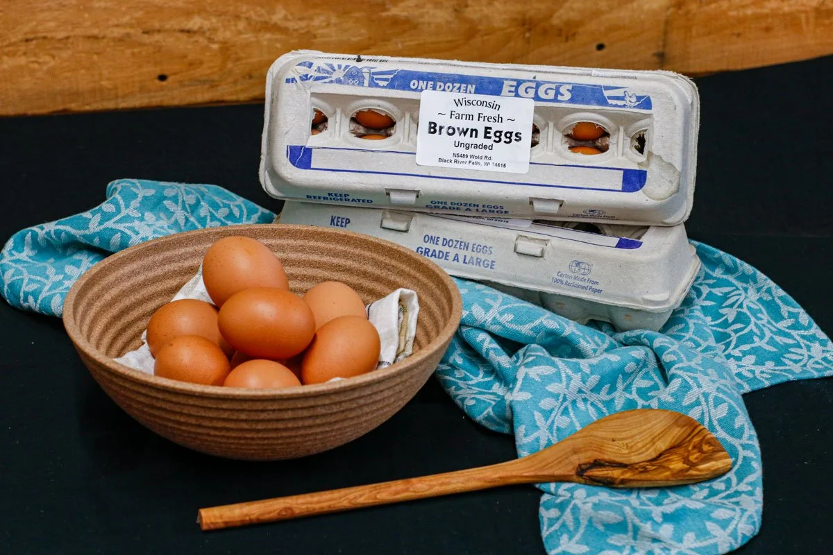 A carton of brown eggs with a label that reads "Wisconsin Farm Fresh Brown Eggs Ungraded," a brown bowl filled with more brown eggs, and a wooden spoon placed on a blue patterned cloth on a black surface.