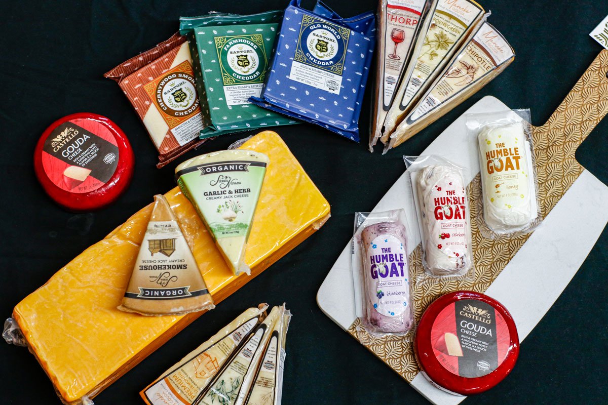 A variety of cheeses, cheese spreads, and goat cheeses displayed on a table, including cheddar, gouda, garlic herb, and flavored goat cheese with different packaging and flavors.