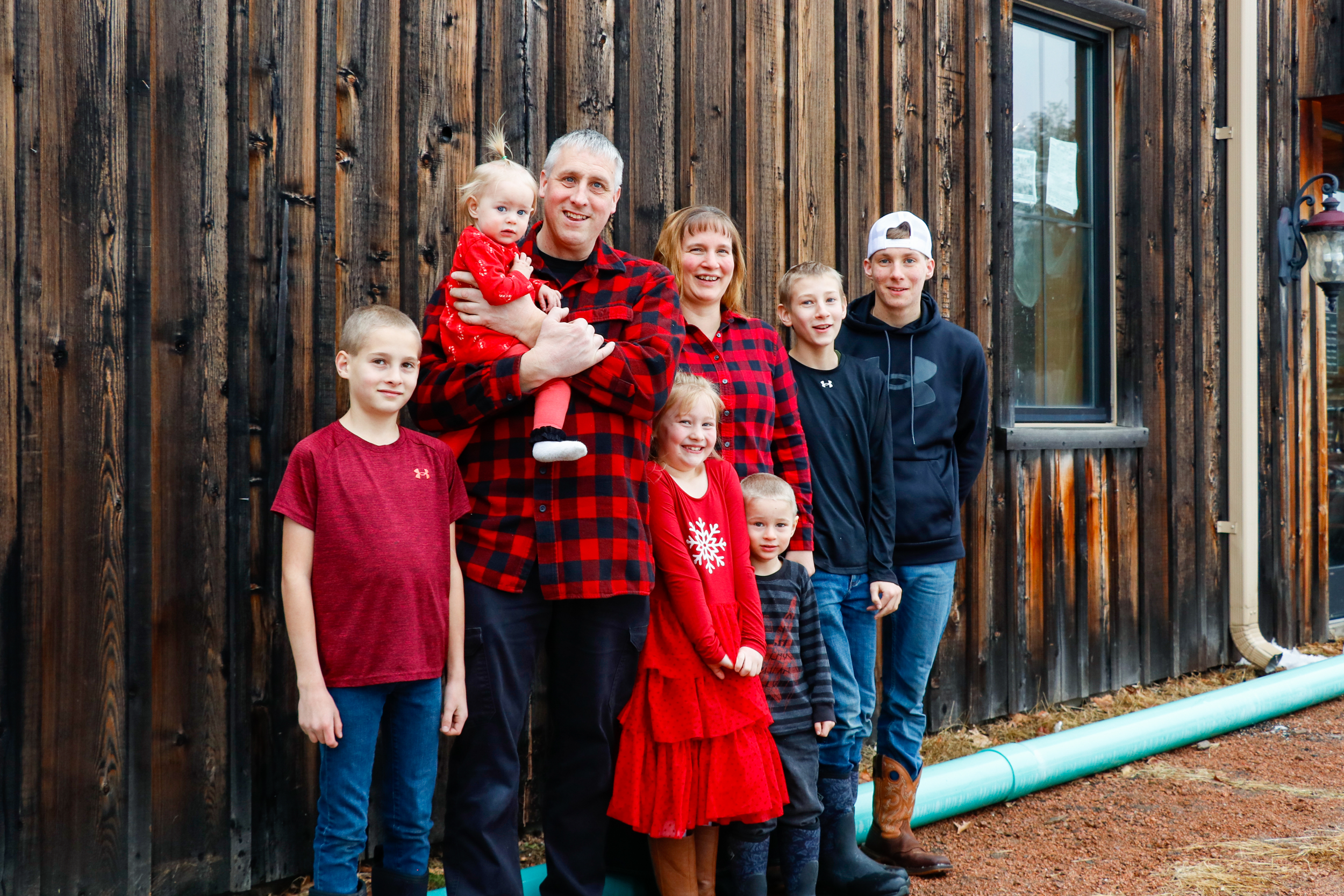 Family of eight standing outdoors in front of a wooden wall, dressed in red and black plaid and casual clothing, smiling for a group photo.