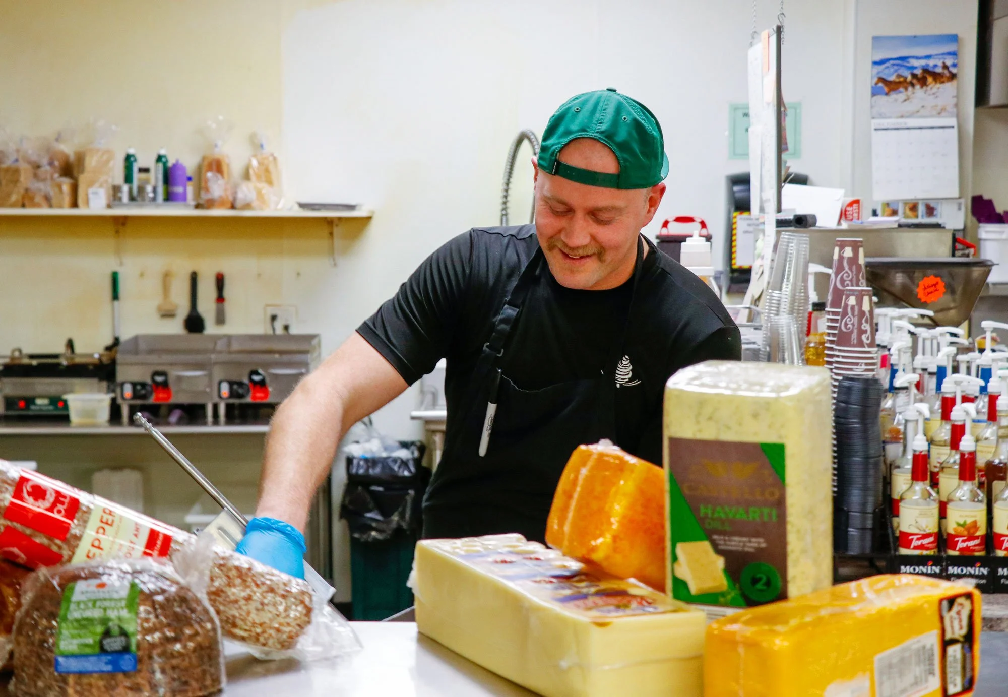 A smiling man wearing a green cap and black apron working behind a cheese counter in a store, surrounded by various types of cheeses and condiments.