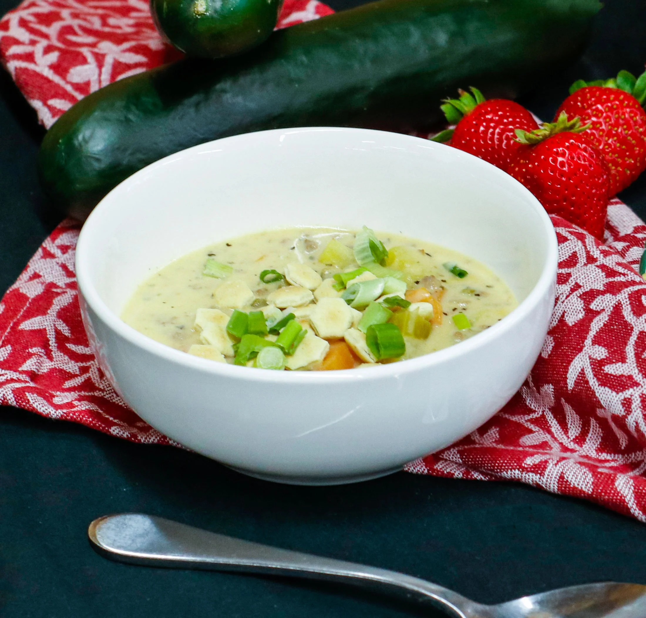 Bowl of potato soup garnished with chopped green onions on a red and white patterned cloth, with cucumbers and strawberries in the background.