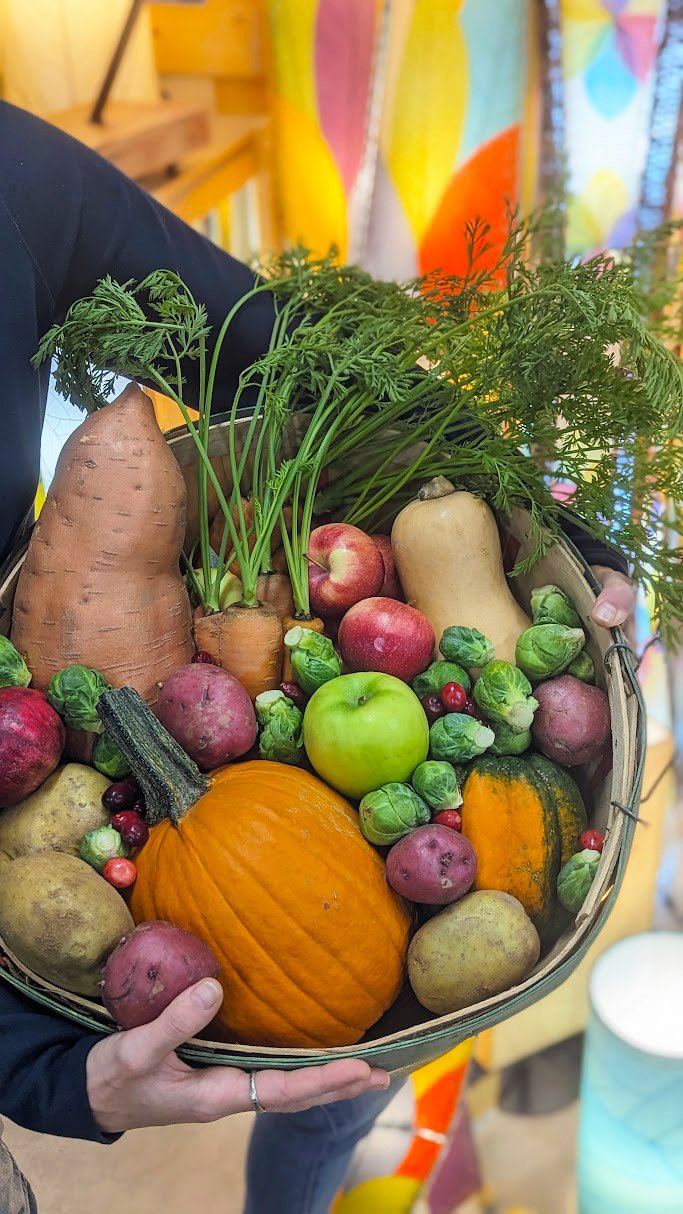 Farm fresh local fall produce in a basket featuring sweet potatoes, pumpkin, squash, Brussel sprouts, apples and potatoes.
