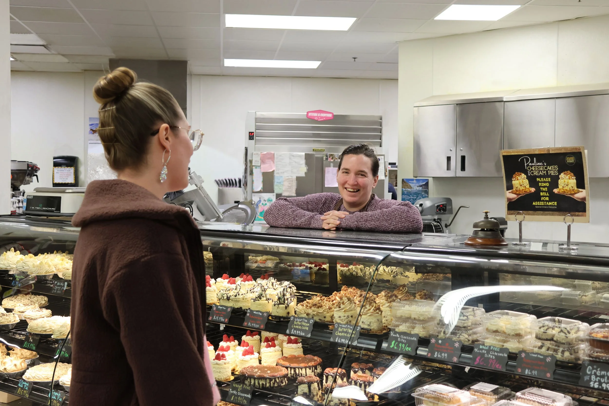 A woman with glasses and earrings talking to a smiling bakery clerk behind a glass display of assorted cakes and pies in a bakery store.