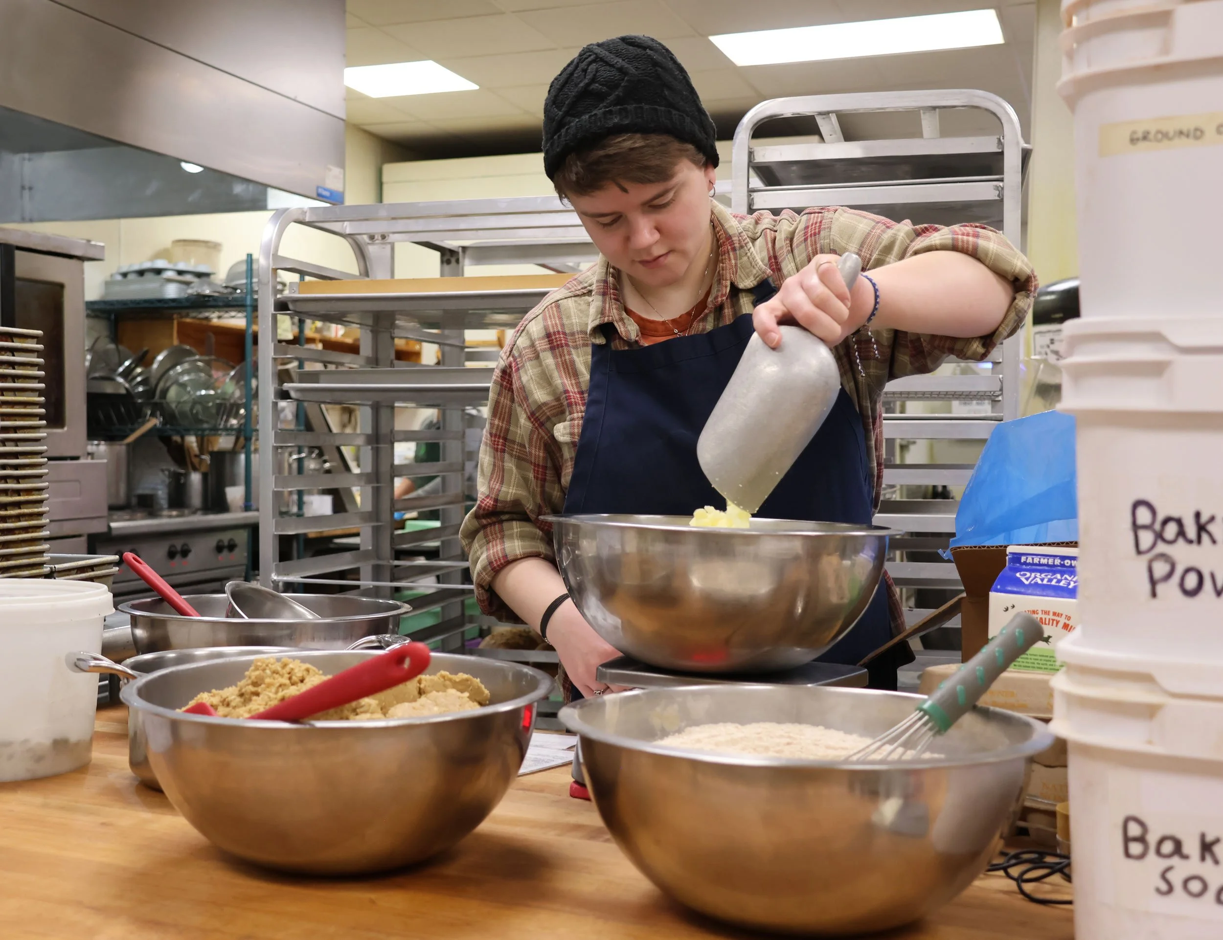 A person pouring ingredients into a large mixing bowl in a commercial kitchen surrounded by metal bowls, kitchen utensils, and baking supplies.