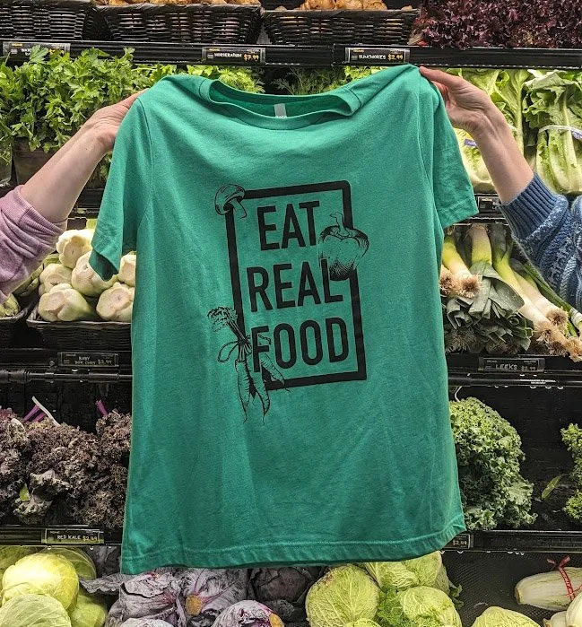 Eat Real Food T-shirt being held up in front of the produce coolers.