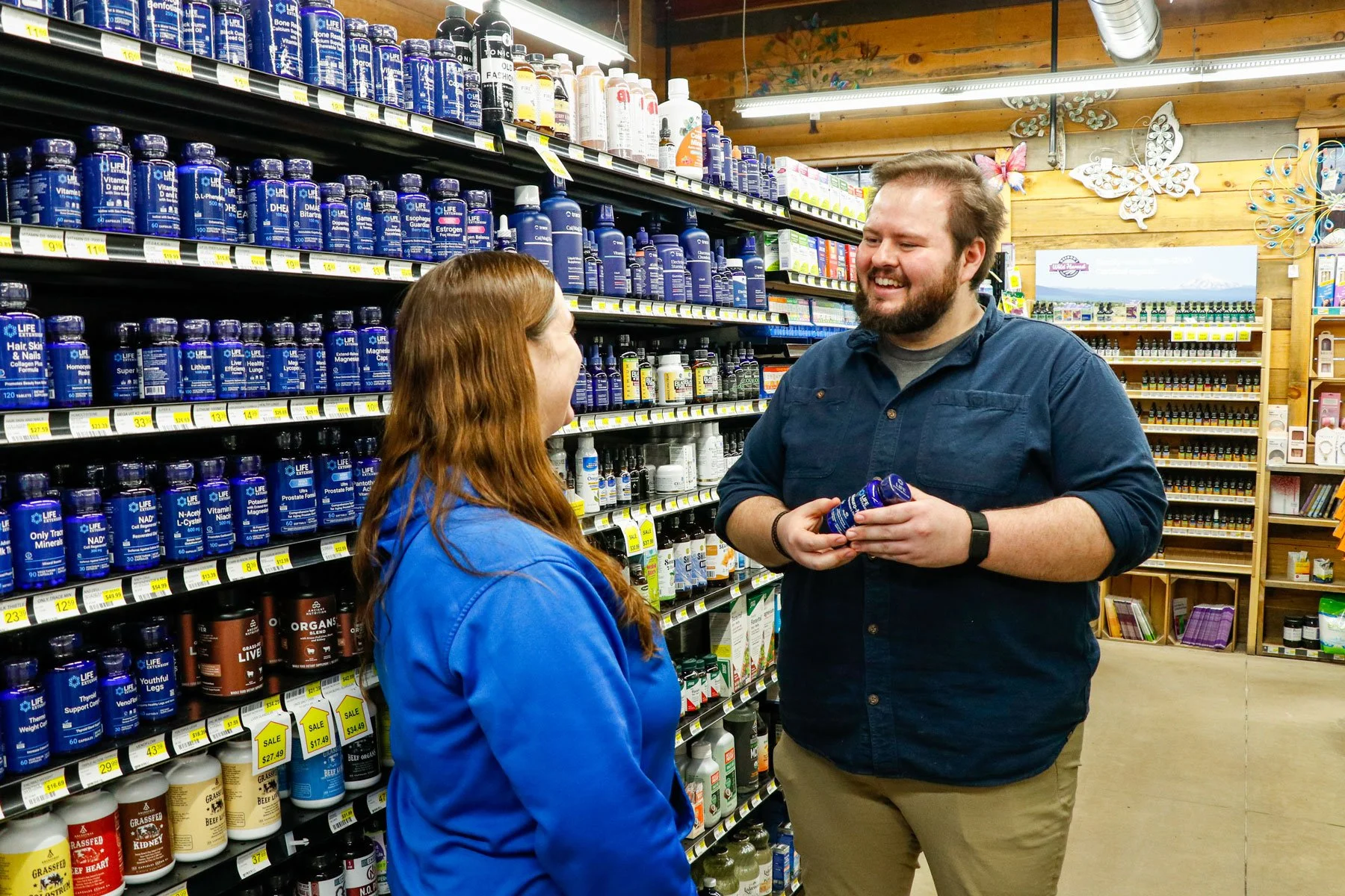 A man and woman talking in a supplement store aisle. The man is smiling and holding a bottle of vitamins. The woman is wearing a blue jacket, and the man is dressed in a dark shirt and khaki pants. The shelves are stocked with various supplement bottles.
