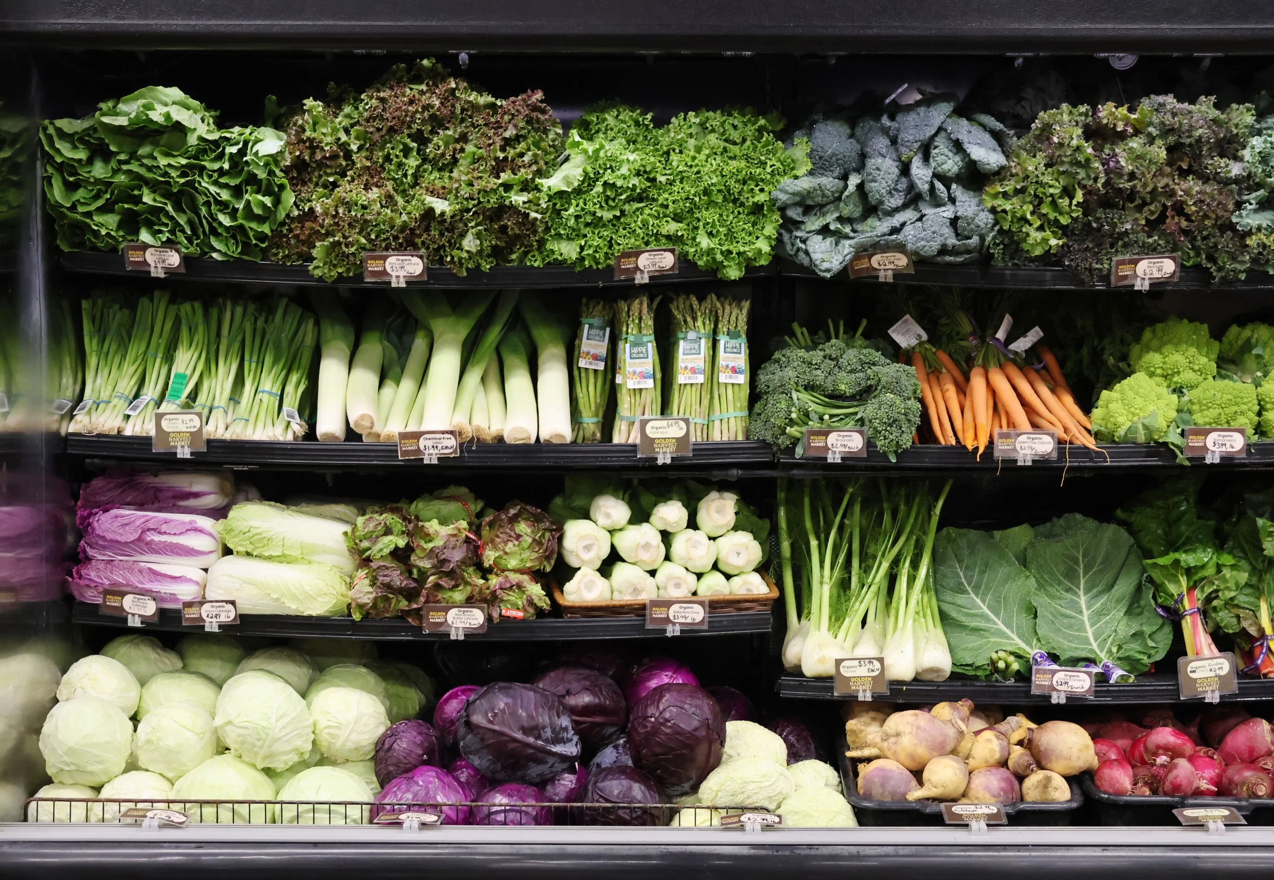 A display of various fresh vegetables on the shelves at a grocery store, including lettuce, kale, asparagus, broccoli, carrots, cauliflower, red cabbage, radishes, and other greens.