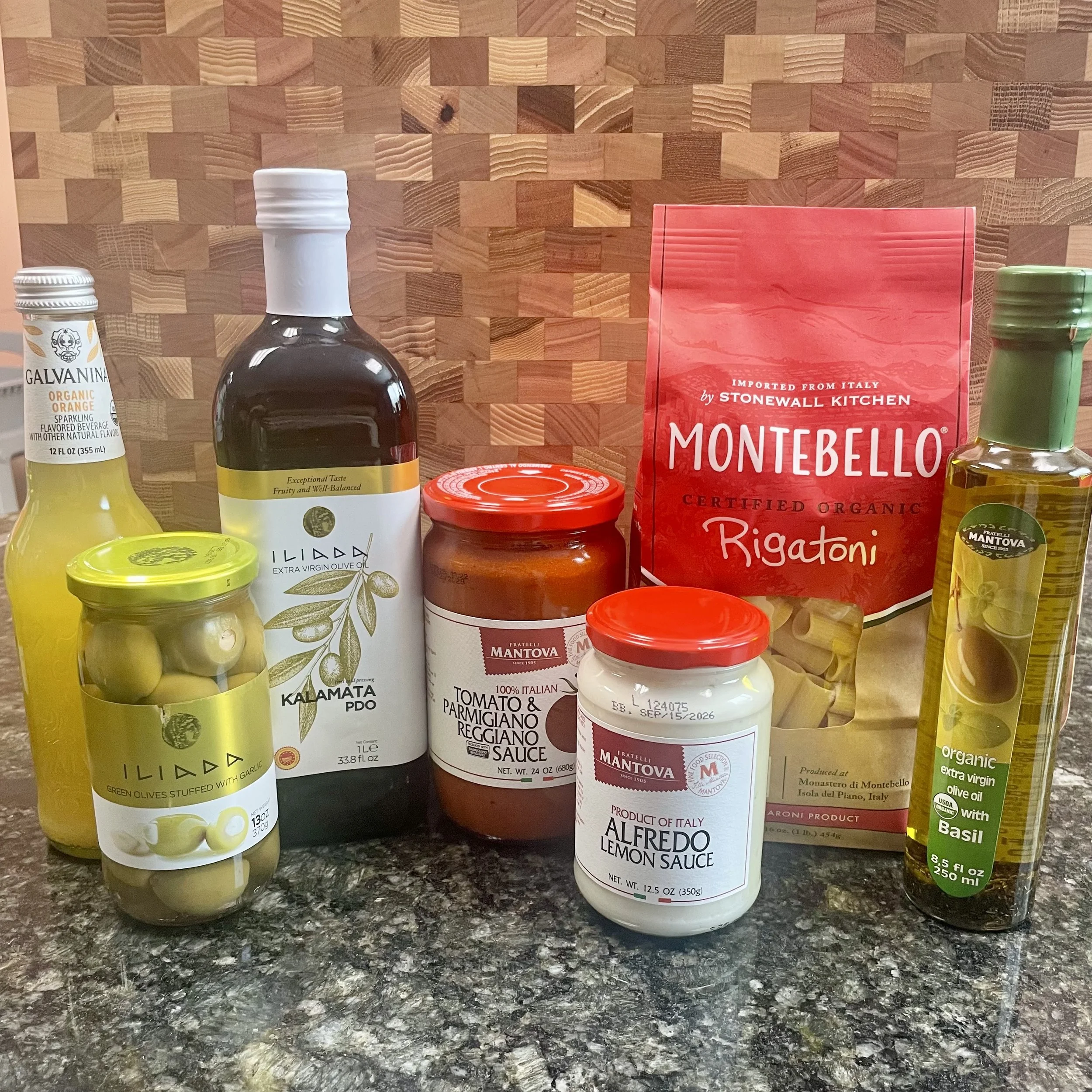 Specialty imported grocery items featuring garlic stuffed olives, rigatoni pasta, Alfredo lemon pasta sauce, extra virgin olive oil, and orange Italian soda.
