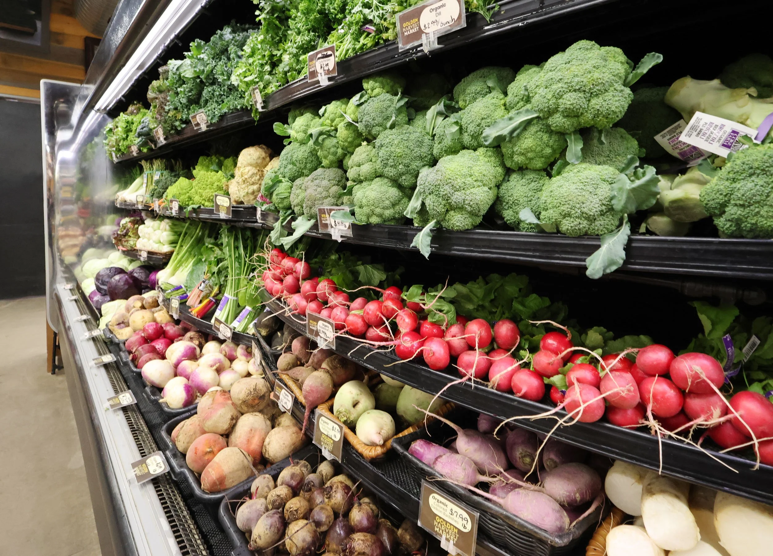 Fresh vegetables including broccoli, radishes, turnips, celery, cauliflower, and leafy greens on display in a grocery store produce section.