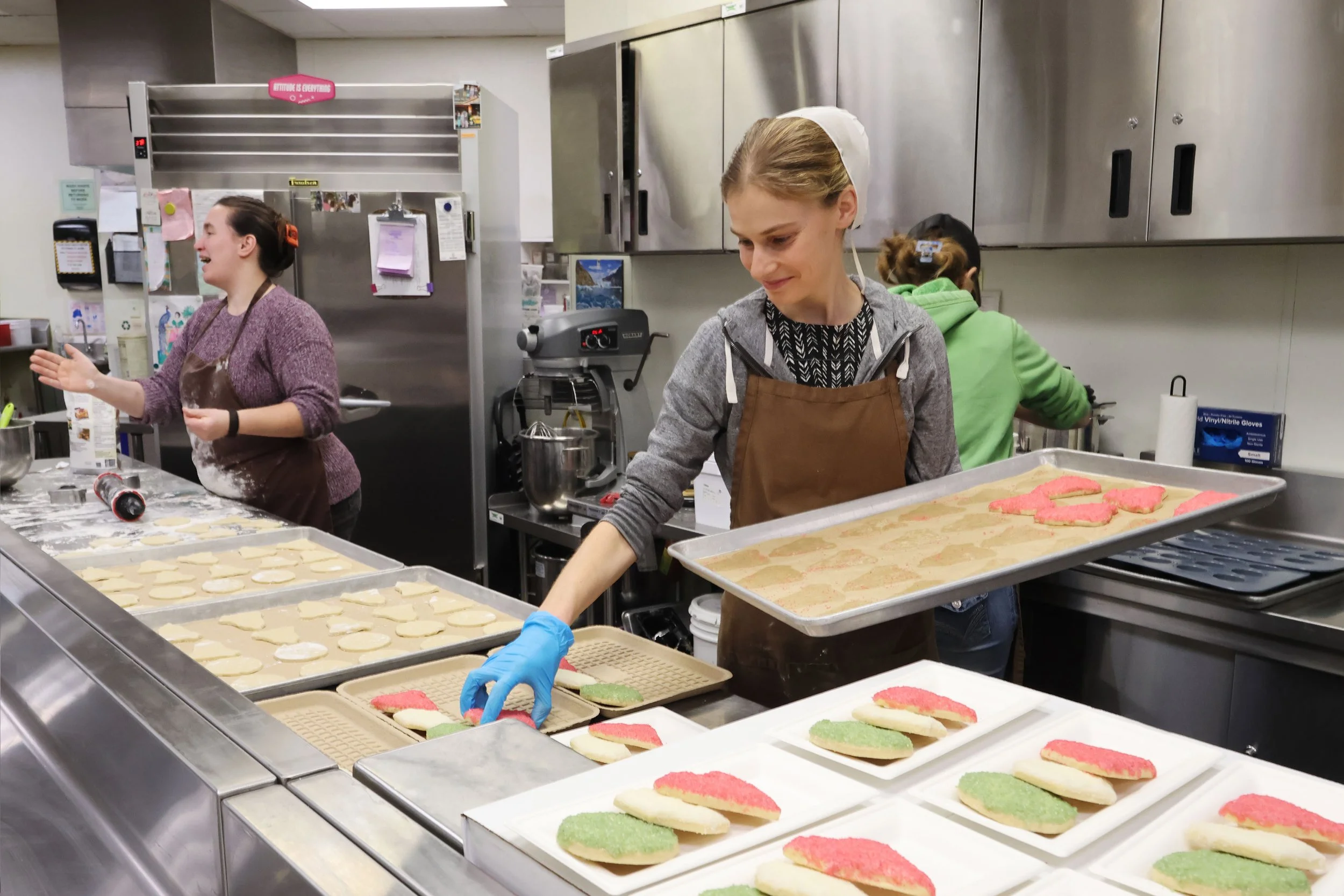 Three women in a commercial kitchen preparing decorated cookies. One woman in the foreground is wearing a brown apron and blue gloves, placing cookies on trays. The woman on the left is smiling and gesturing, while the woman in the background is working at a countertop.