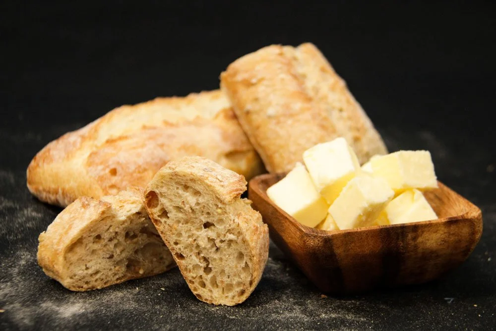 Pieces of sliced French bread and a small wooden bowl filled with butter on a black surface.