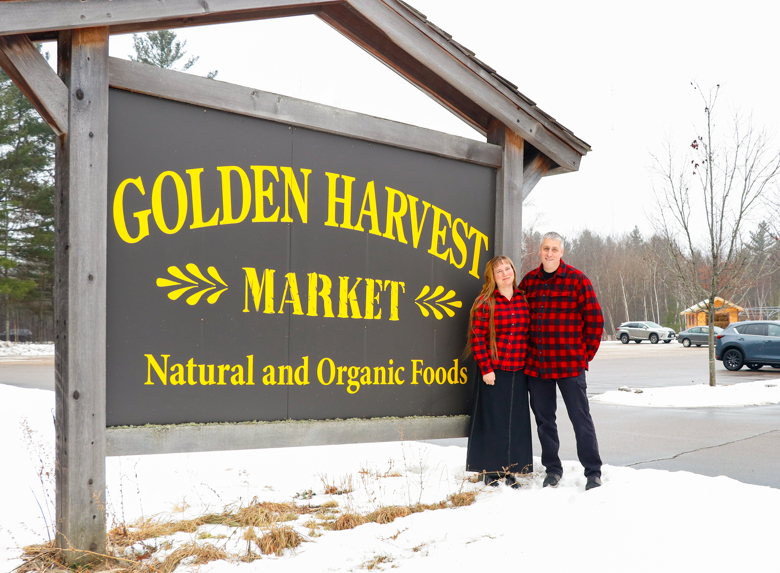 A couple standing outside near a sign that reads 'Golden Harvest Market, Natural and Organic Foods,' on a snowy day, wearing matching red and black checkered shirts.