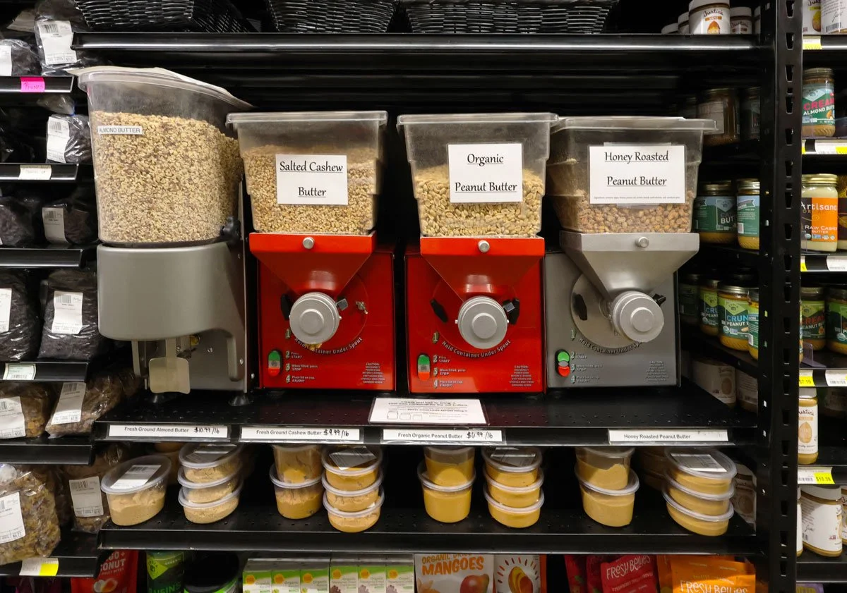 Three bulk bins containing almond butter, salted cashew butter, and organic peanut butter, with labeled signs; small containers of peanut butter and other products on lower shelves.