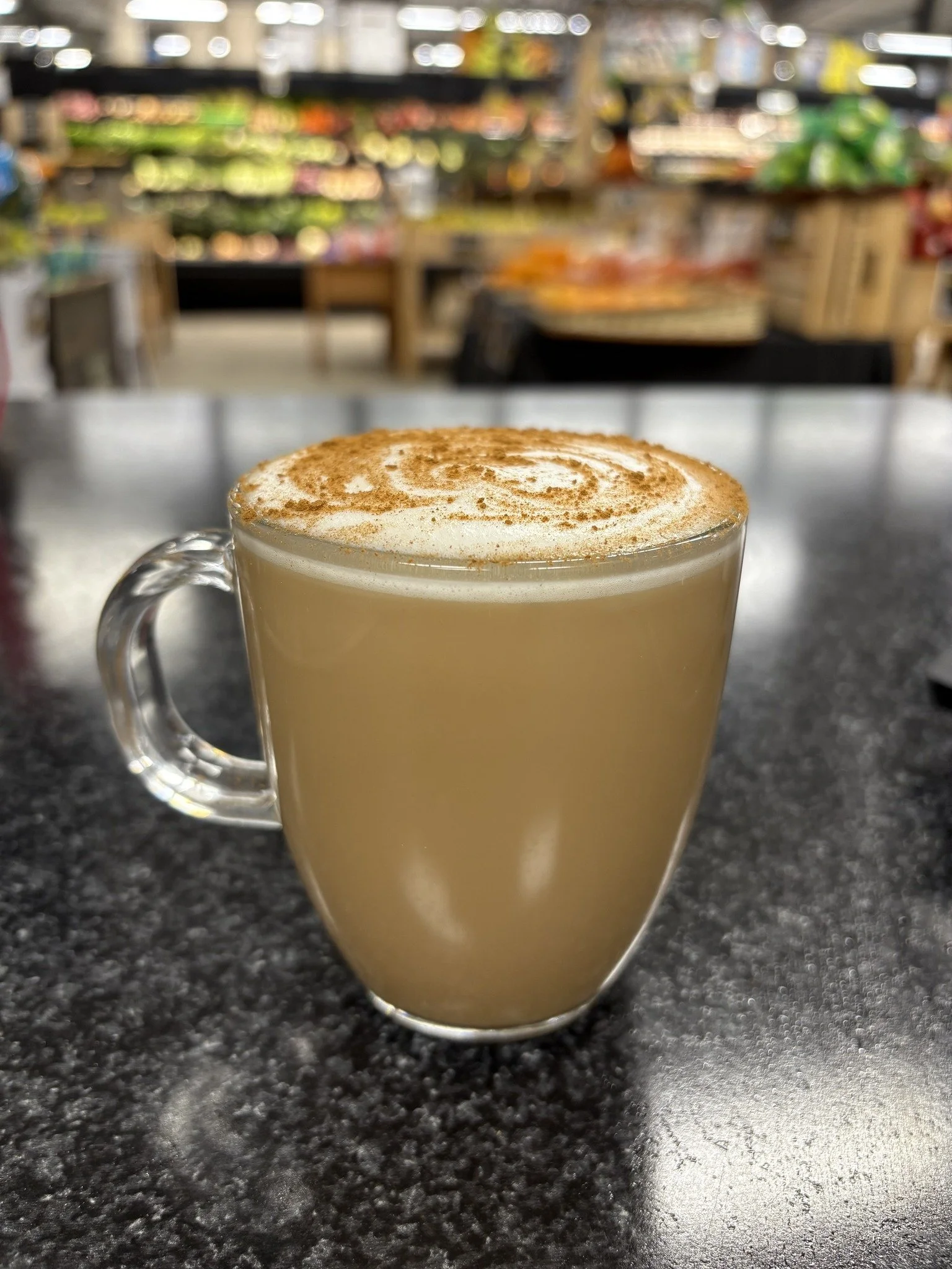A clear glass mug filled with a latte topped with cinnamon, placed on a black granite table in a grocery store or café setting.