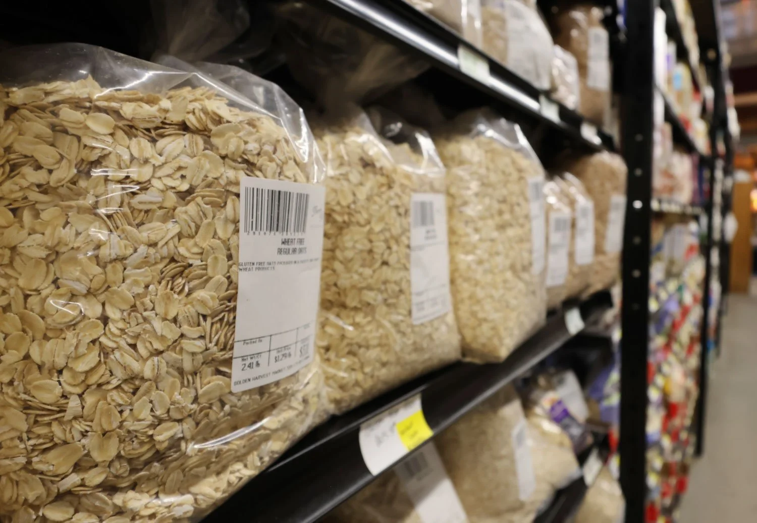 Bulk section of a grocery store featuring clear plastic bags of oats on a black metal shelf, with a focus on a bag of rolled oats in the foreground.
