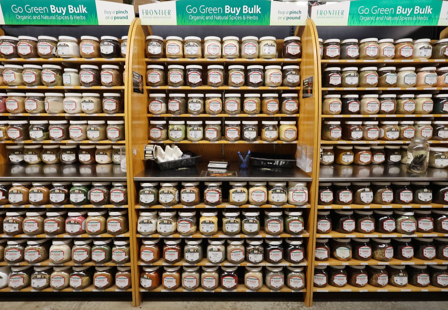 Wall of spice jars at a grocery store, organized on wooden shelves with a variety of spices and herbs. Green banners above promote buying organic and natural spices in bulk.