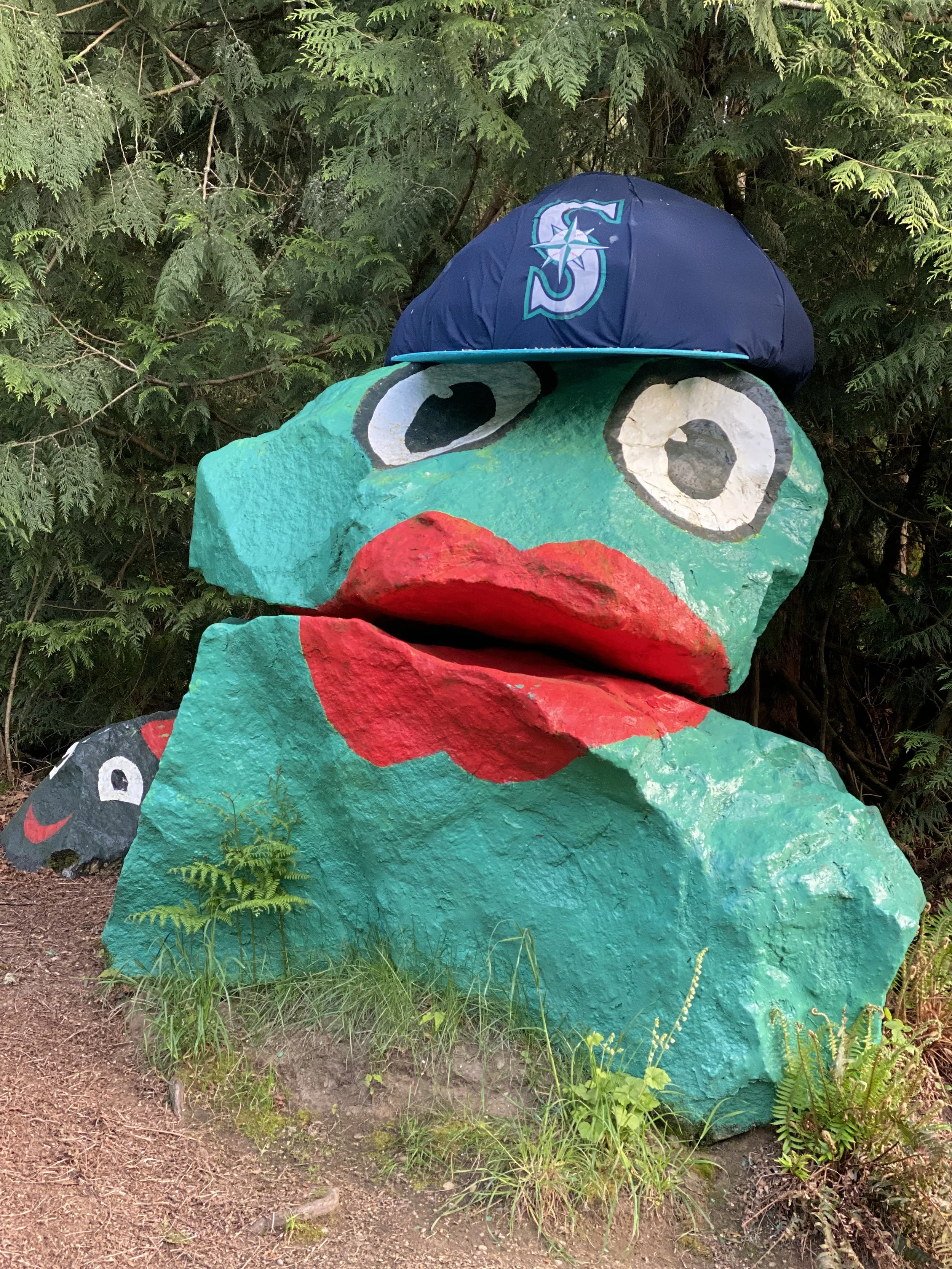 A large, colorful painted Frog Rock sculpture with big eyes and red curlicue lips, wearing a Seattle Mariners baseball cap, is situated outdoors against green foliage.