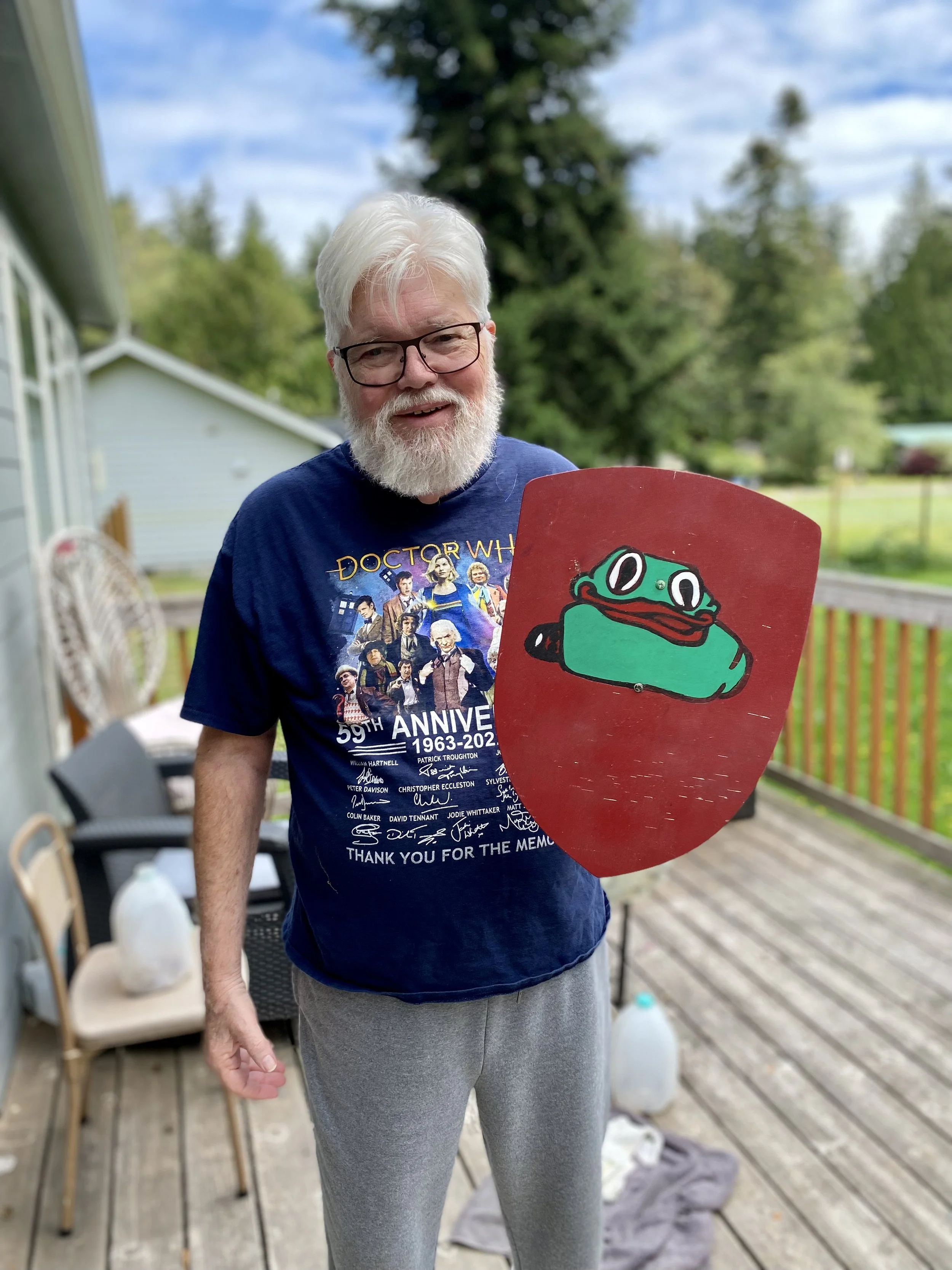 A smiling elderly man with gray hair and beard, wearing glasses and a blue graphic T-shirt, stands on a wooden deck holding a shield with a cartoon frog design. The background shows trees, a blue house, and outdoor furniture.