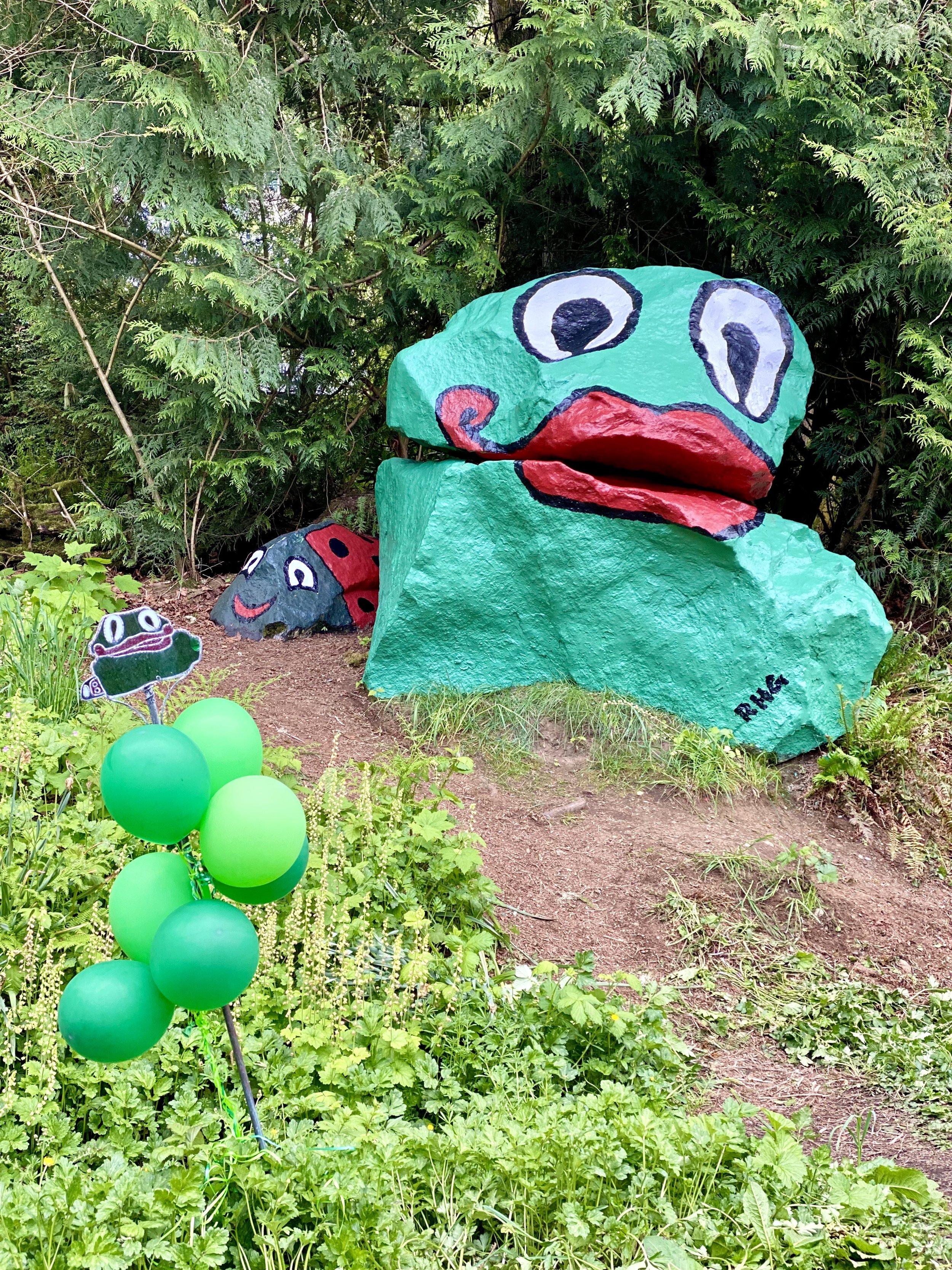 A large, cartoonish painted Frog Rock sculptures, and a small painted ladybug rock, set outdoors among green foliage and grass, with some green balloons in the foreground.