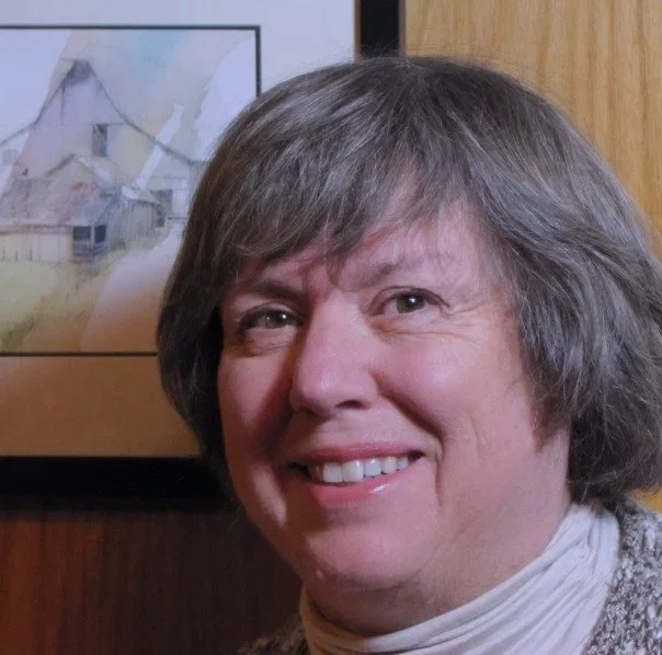 Close-up photo of smiling woman with short gray hair, wearing a white turtleneck, indoors with framed artwork in the background.