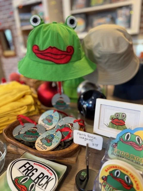 Green bucket hat with frog face and eyes, displayed on a store shelf alongside other hats, magnets, and frog-themed merchandise.