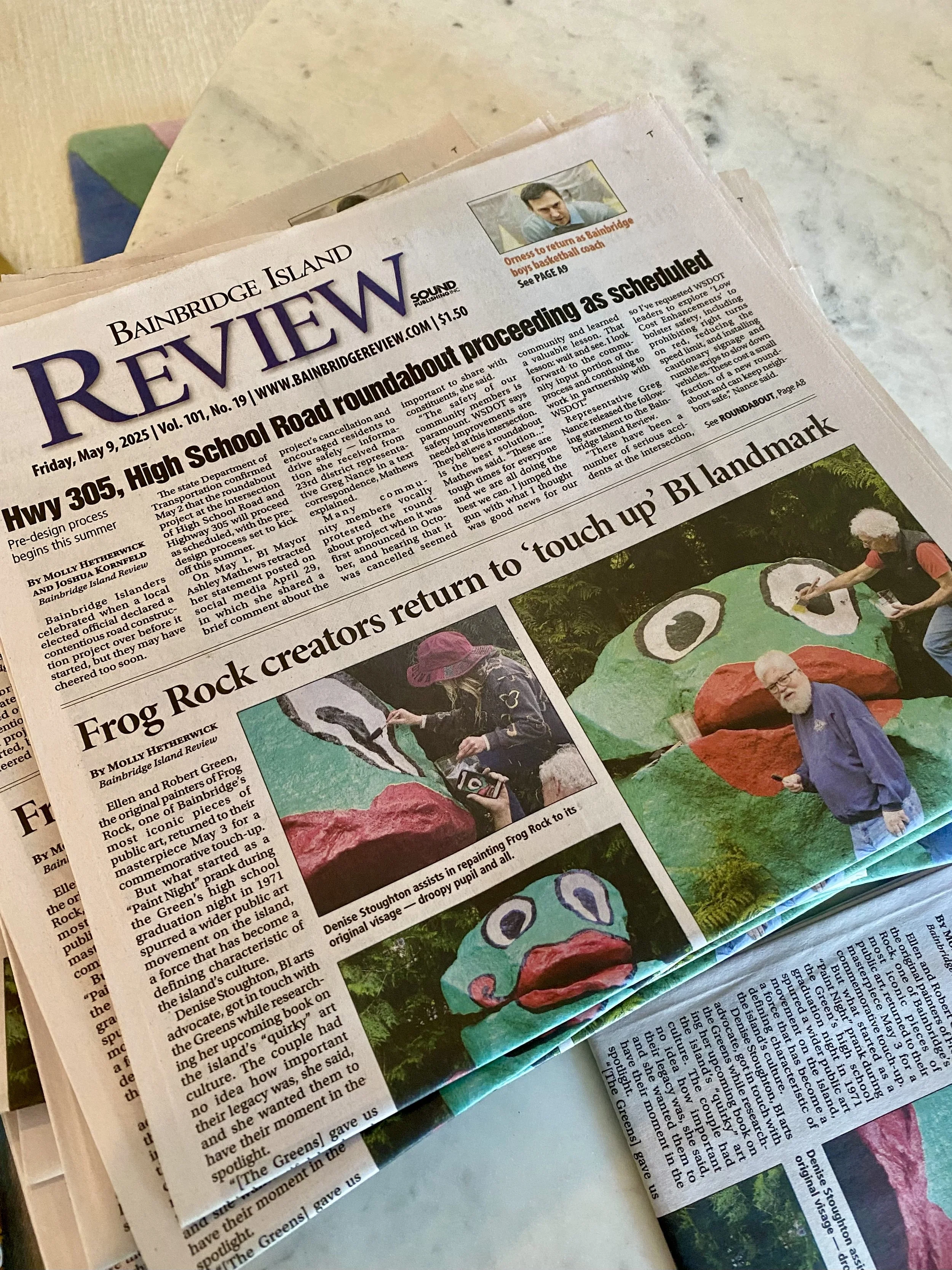 A newspaper page on a table, featuring articles about high school roundabout and Frog Rock art projects, with photos of people working on colorful frog sculptures.