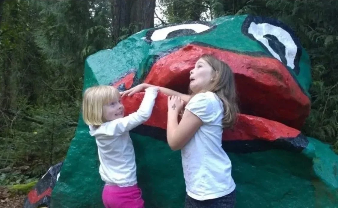 Two young girls are playing in front of a large, colorful, painted Frog Rock in a wooded outdoor area.