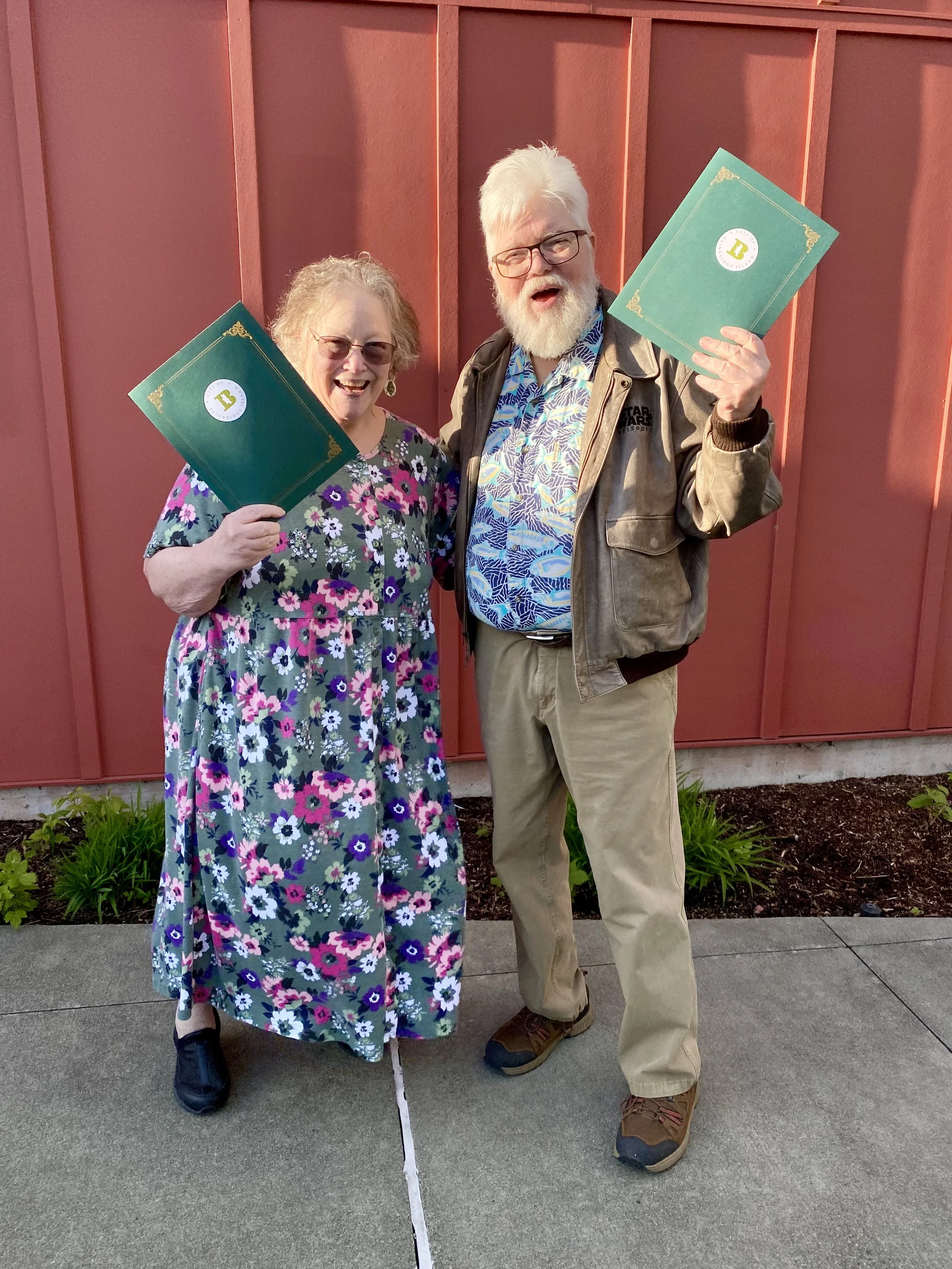 Happy woman and man, Ellen & Bob Green, holding green folders, standing outdoors in front of a red wooden wall, smiling and celebrating.