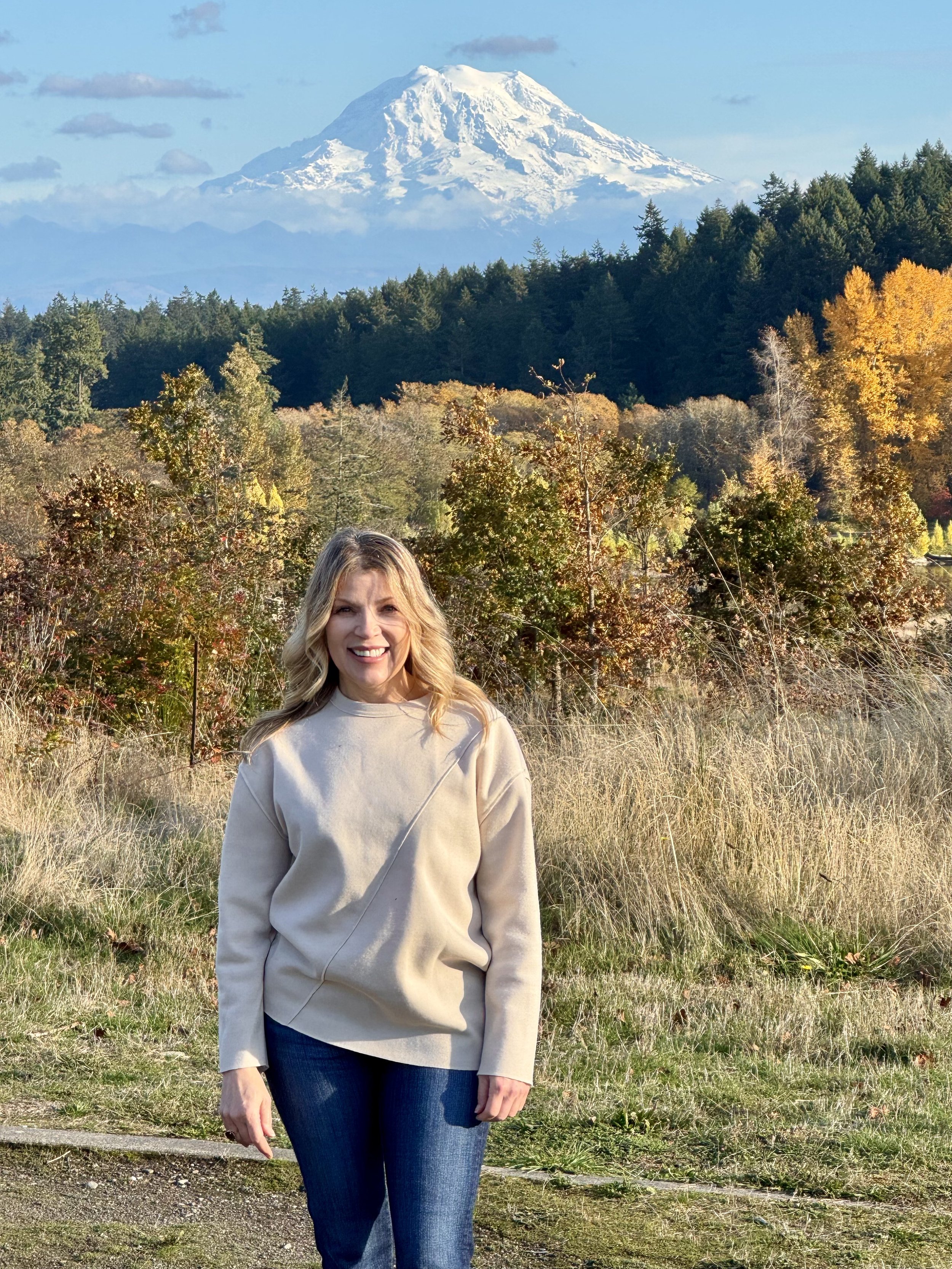 A smiling woman with blonde hair in casual clothing standing outdoors with a mountainous landscape, including snow-capped Mount Rainier in the background, surrounded by trees and autumn foliage.