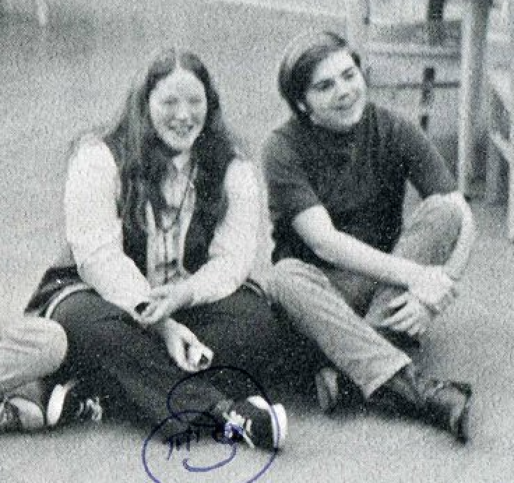 Two teenagers sitting on the floor, smiling, in a room with shelves or storage in the background. One has long hair and the other has short hair.