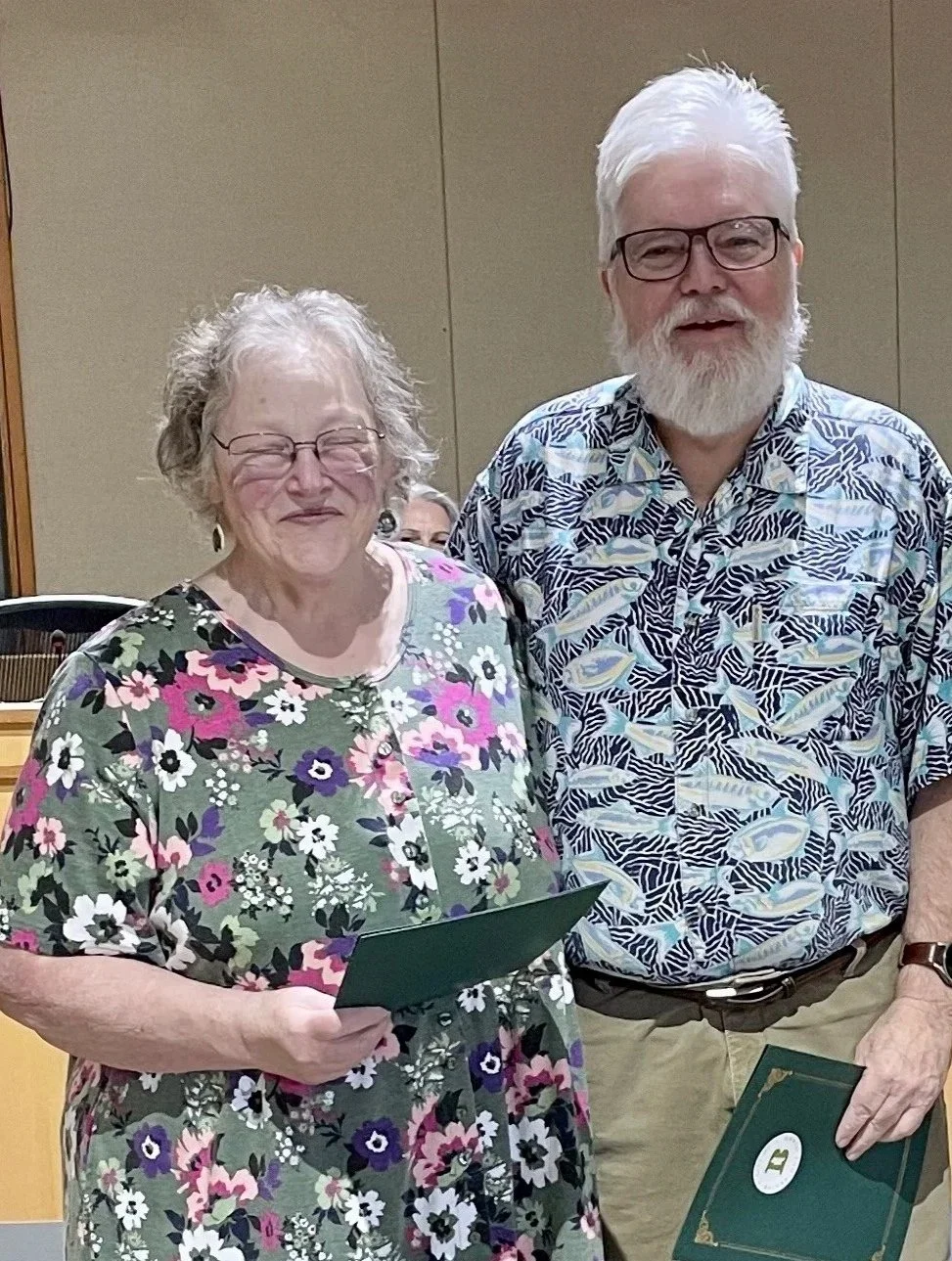 An elderly woman with gray, curly hair and glasses, wearing a floral dress, holding a green paper, standing next to an elderly man with white hair and beard, glasses, wearing a patterned Hawaiian shirt, holding a green folder, smiling indoors.