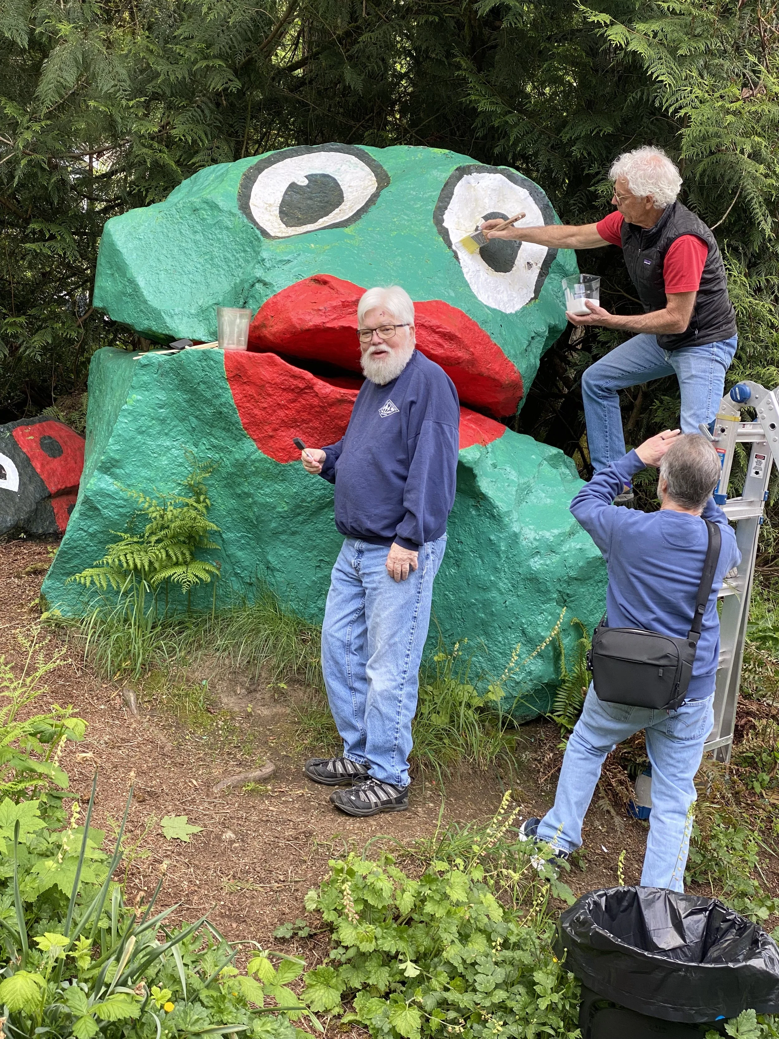 Three men painting and working on a large, colorful natural frog rock sculpture outdoors, with one man in front looking at the camera and two others painting the sculpture.