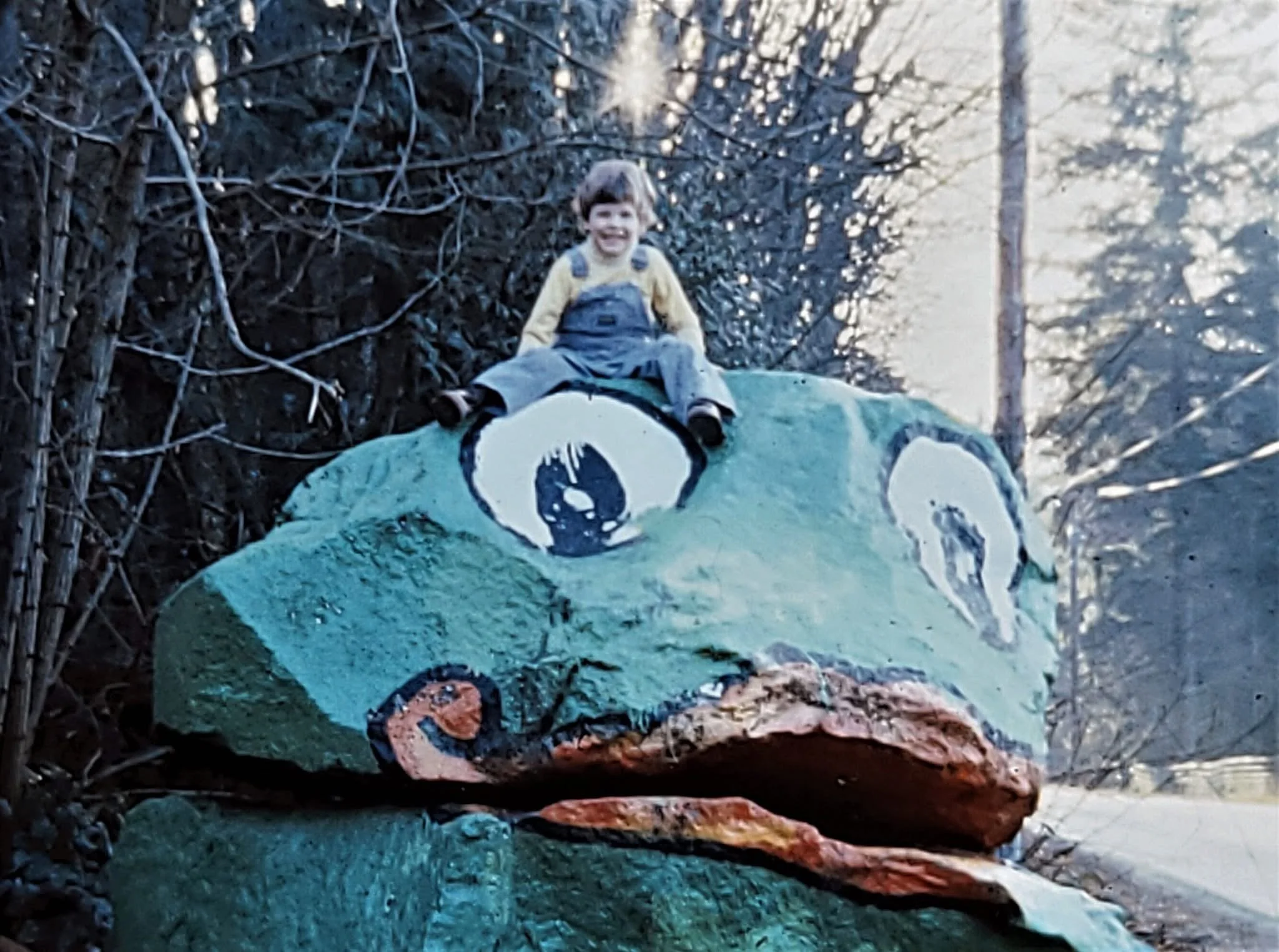 A child sitting on a painted rock sculpture of a frog in an outdoor setting.