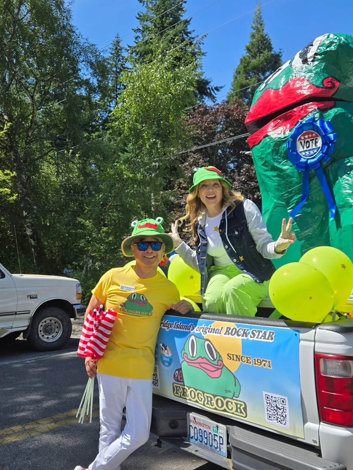 Two women dressed in frog-themed costumes posing with a large frog-shaped float during a parade. One woman is sitting on the float and the other is standing beside it, both smiling. The float has balloons and a banner that reads 'Frogrock,' featuring a frog cartoon.