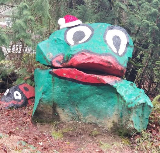 Large painted rock sculpture resembling a frog with big eyes, a red Santa hat, and a wide mouth, situated outdoors with greenery around.