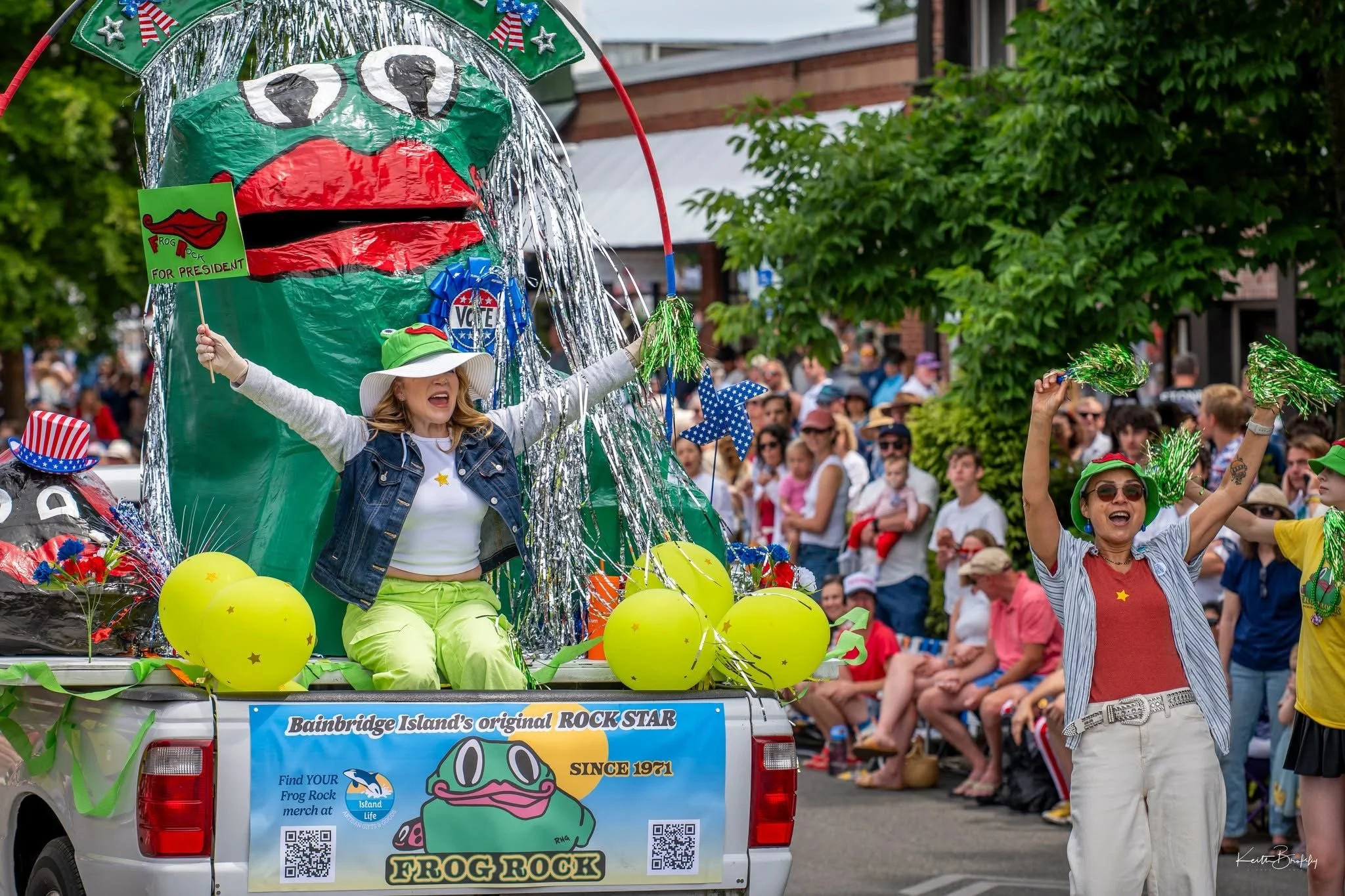 People on a parade float decorated with frog-themed decorations, balloons, and patriotic items, while marching and celebrating in a street parade surrounded by spectators.