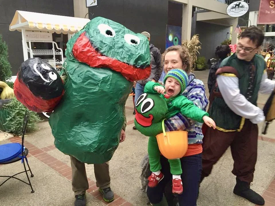 People in costume, including a man dressed as a large green Frog Rock and a crying toddler dressed as a small Frog Rock, being held by a woman at a Halloween street festival on Bainbridge Island.