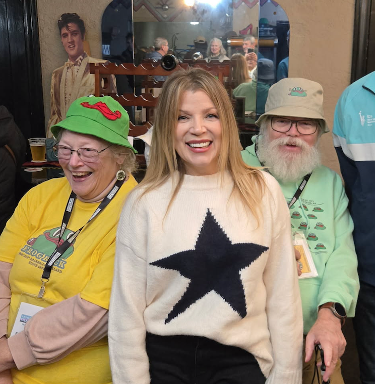 Three smiling people posing for a photo at a social event, with a woman in the center wearing a white sweater with a black star, and an older woman on the left wearing a bright green hat and a yellow shirt, and an older man on the right wearing glasses, a light green shirt, and a beige hat.