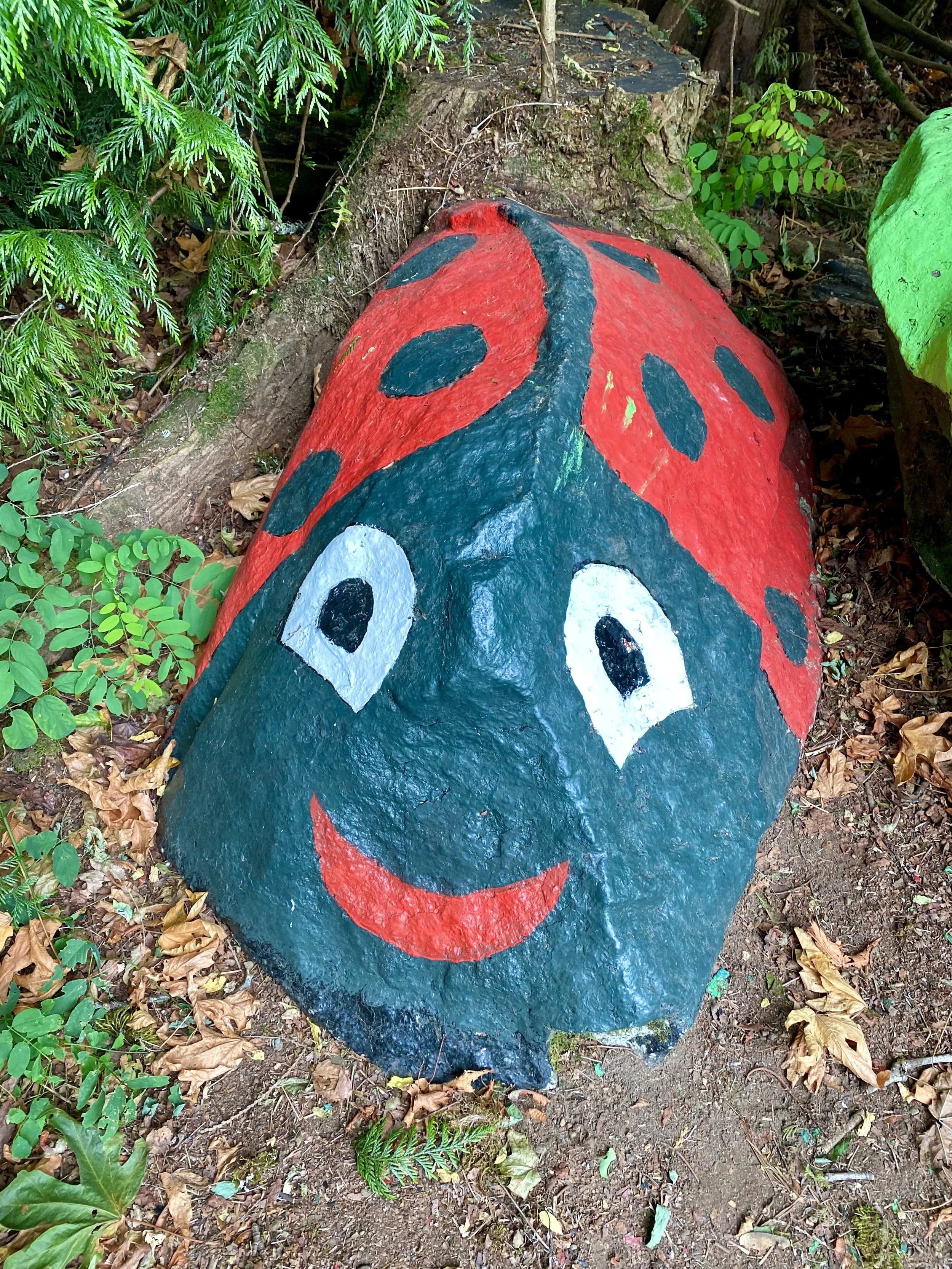 A large rock painted to resemble a smiling ladybug with red and black spots and a smiling face, situated among plants and fallen leaves in a natural outdoor setting.