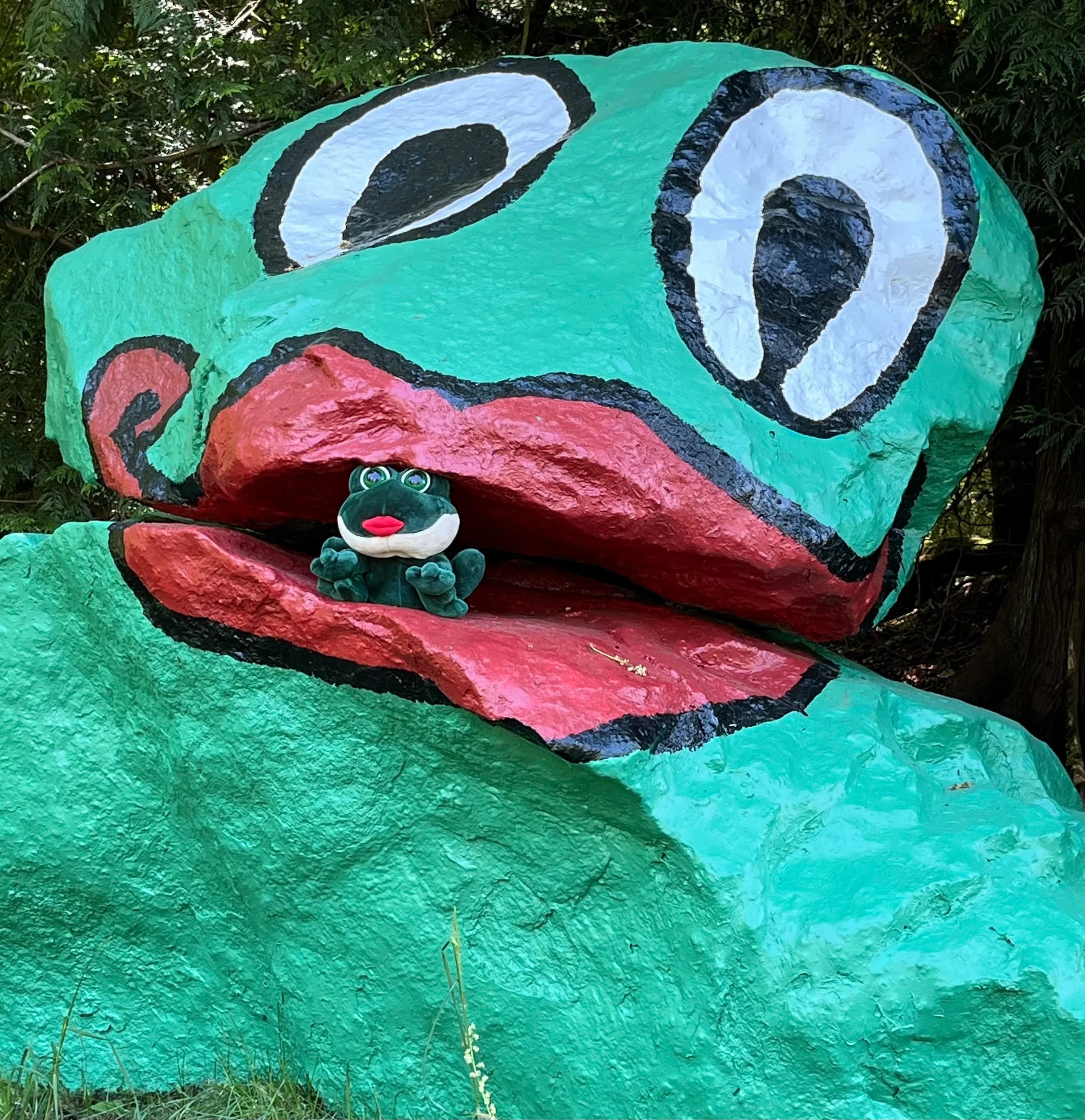 A colorful, cartoonish painted frog rock sculpture with large eyes. A plush toy frog with big eyes, green body, and red lips is positioned in the open mouth of the sculpture.