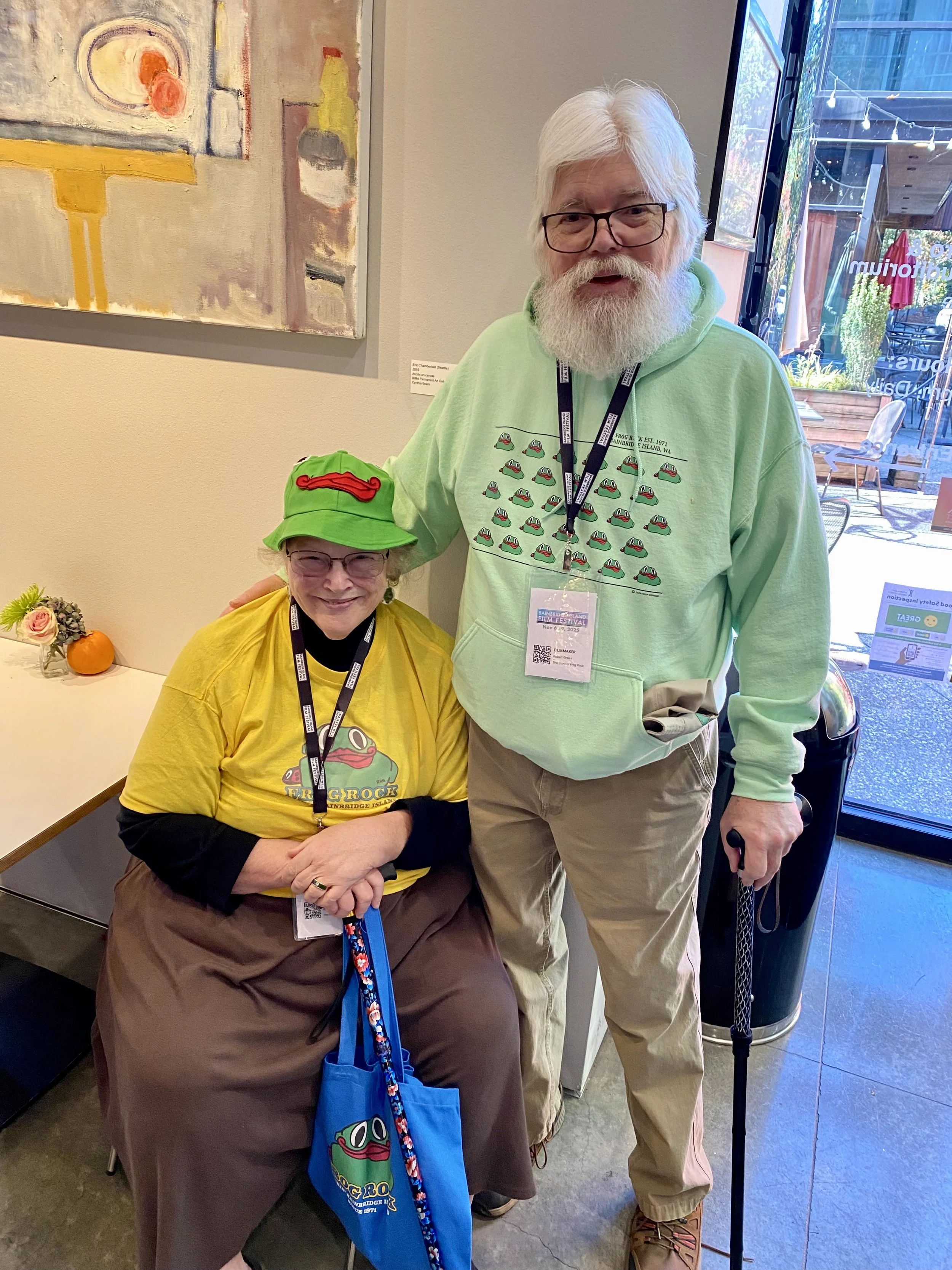 Two people dressed in frog-themed clothing, posing together indoors near an art piece and a window, with a small orange pumpkin and a flower arrangement on the table nearby.