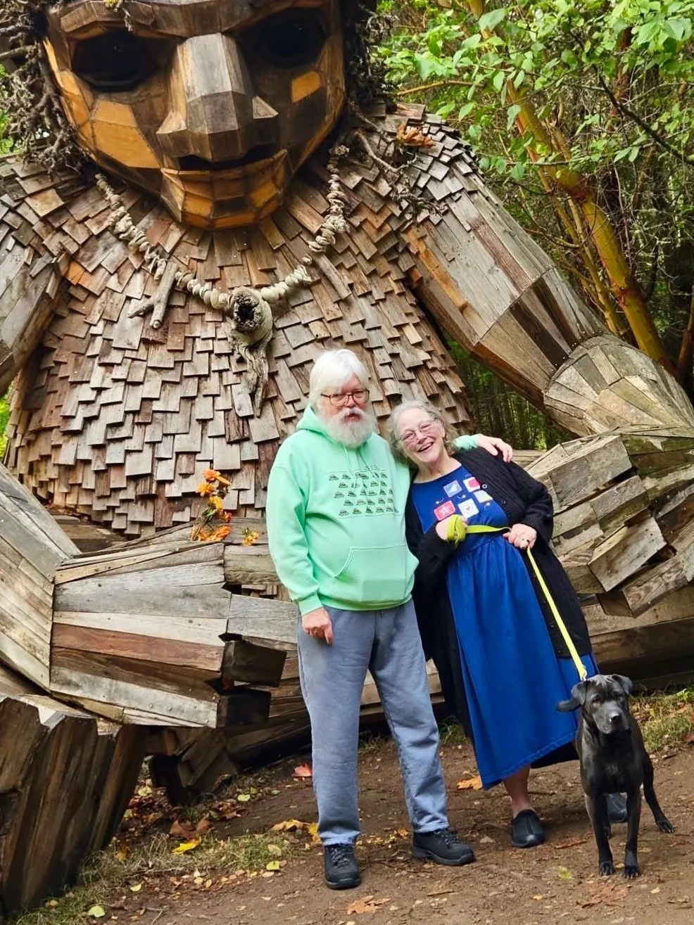 Bob & Ellen Green standing in front of Thomas Dambos' large-scale troll made of recycled wood. Ellen is holding a leash attached to their black dog, Wednesday.