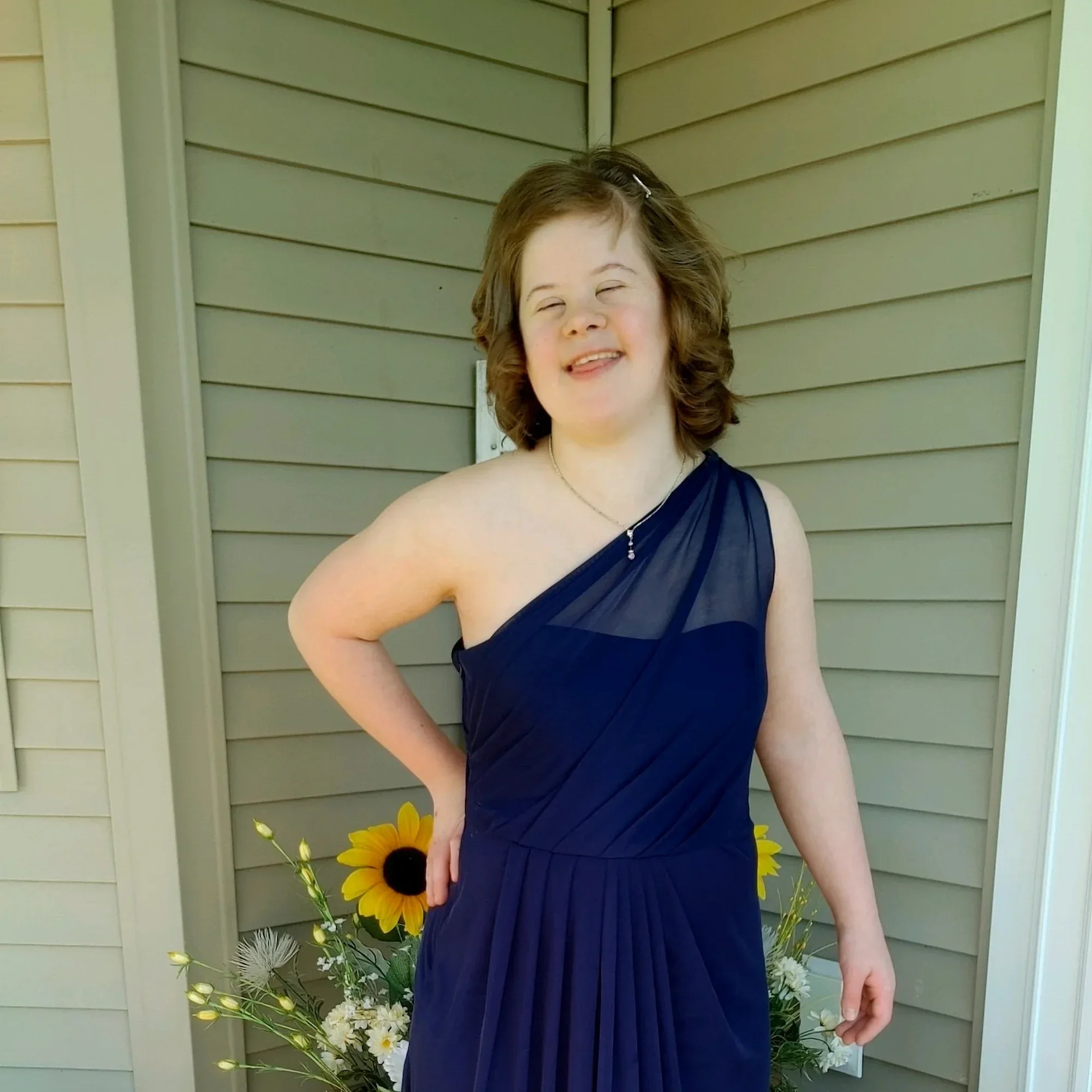 Annaka Paige Werginz, smiling and wearing a one-shoulder navy blue dress, standing outdoors in front of a house with beige siding and a bouquet of sunflowers and white flowers.