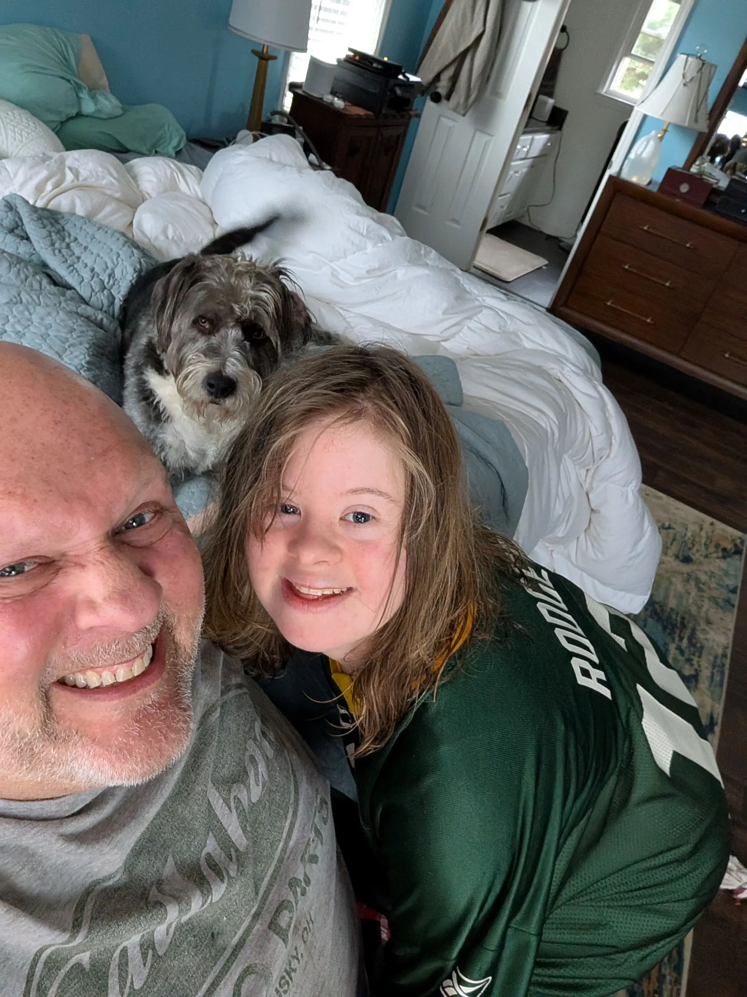 A man, a young girl, and a dog taking a selfie in a bedroom. The man has a wide smile, the girl has her mouth open in a smile, and the dog is lying on the bed looking at the camera.