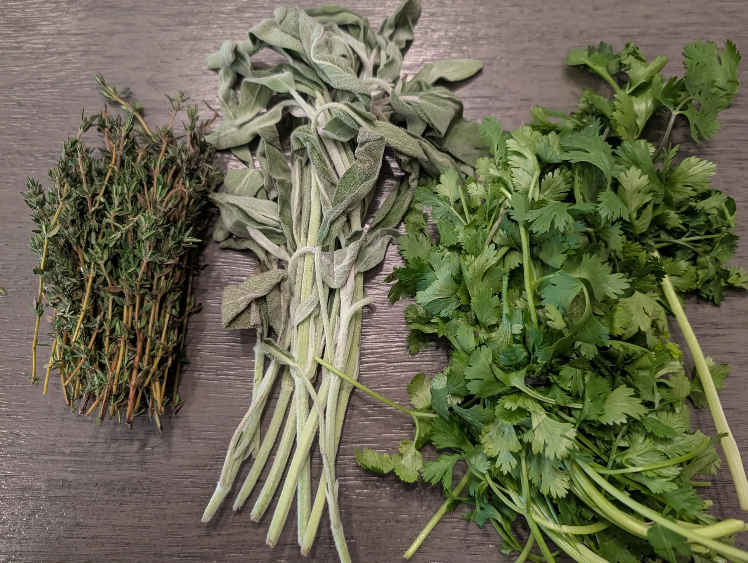 Bundles of fresh herbs including thyme, sage, cilantro, and oregano on a wooden surface.